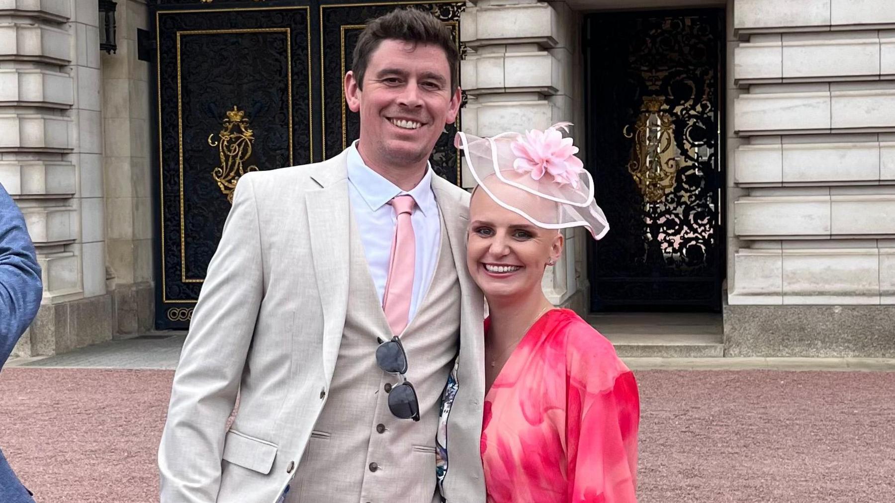A man in a white suit with a pink tie is next to a woman dressed in pink with a pink fascinator on her head. They're both standing close to each other smiling.
