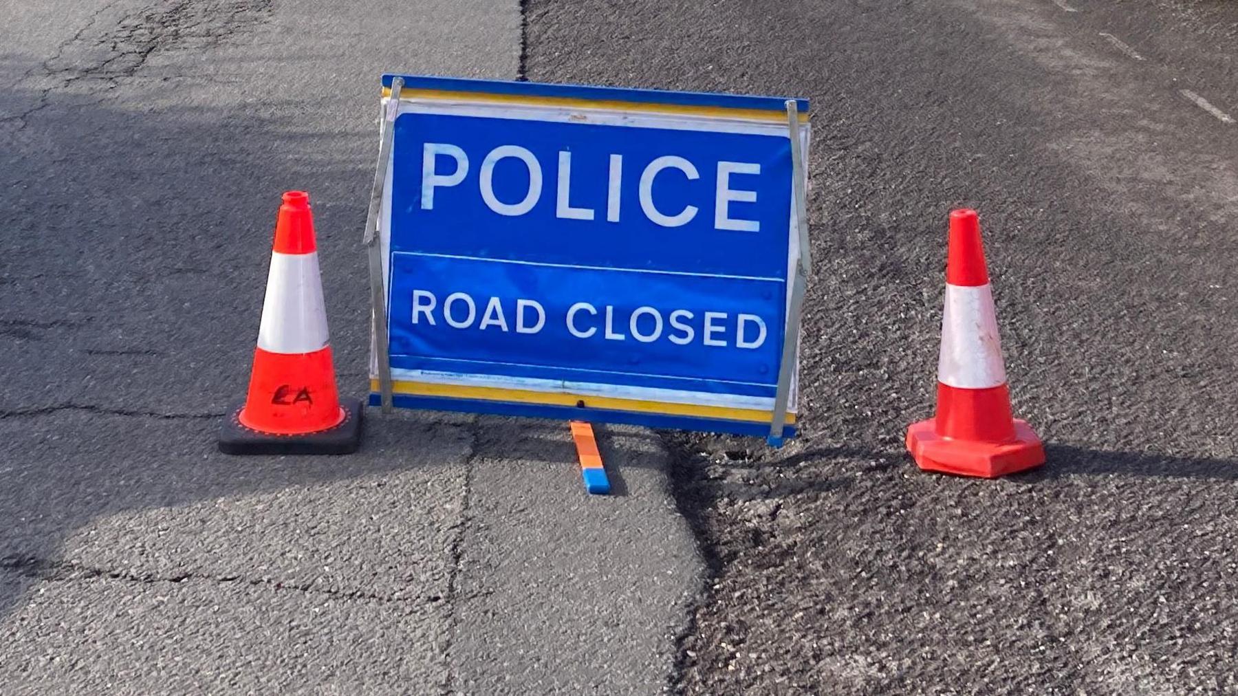 A blue Police Road Closed sign with an orange traffic cone either side on a tarmac road