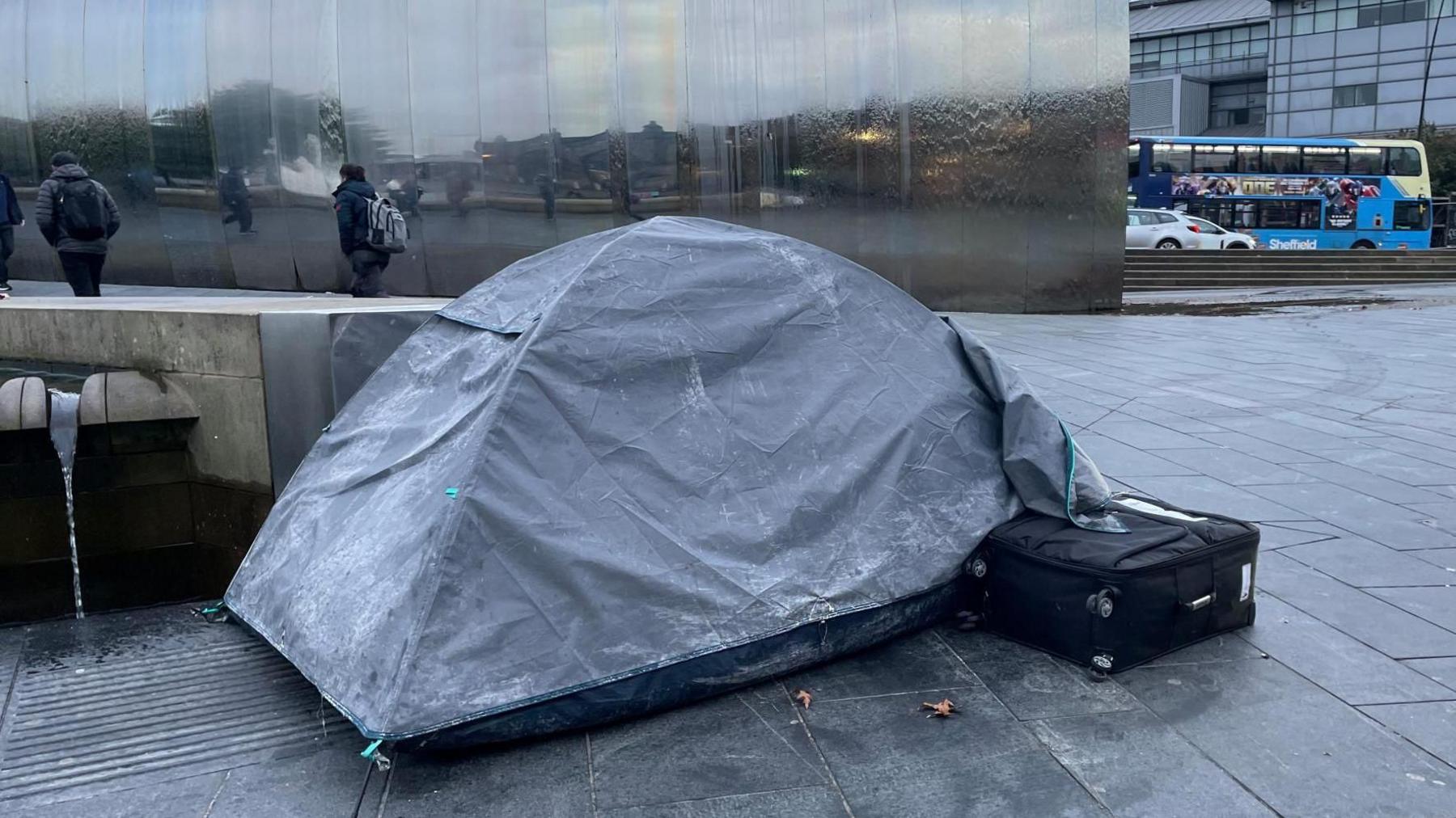 A grey tent on the pavement. People in thick coats walking past.