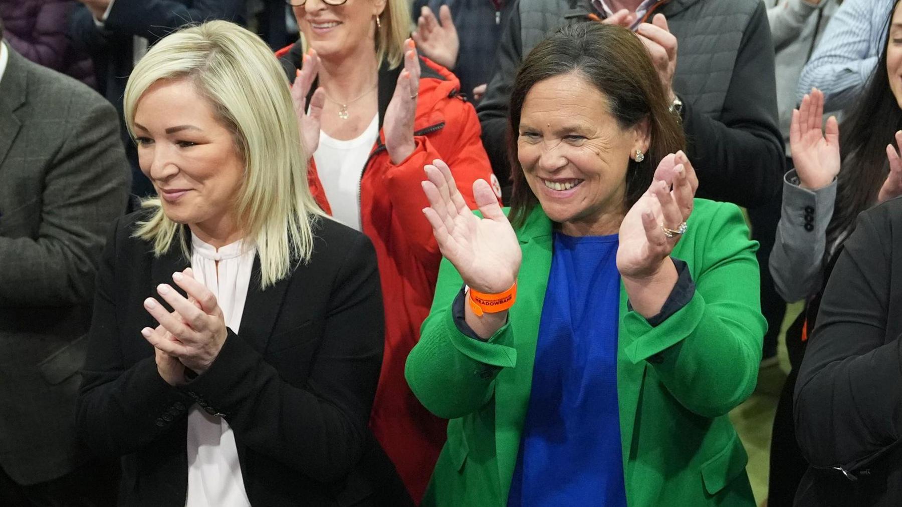 Michelle O'Neill is wearing a black jacket and  white top. She has shoulder length blond hair. Mary Lou McDonald has short brown hair. She's wearing a blue top, with a green jacket. Both women are smiling and clapping their hands. There's a crowd around them.