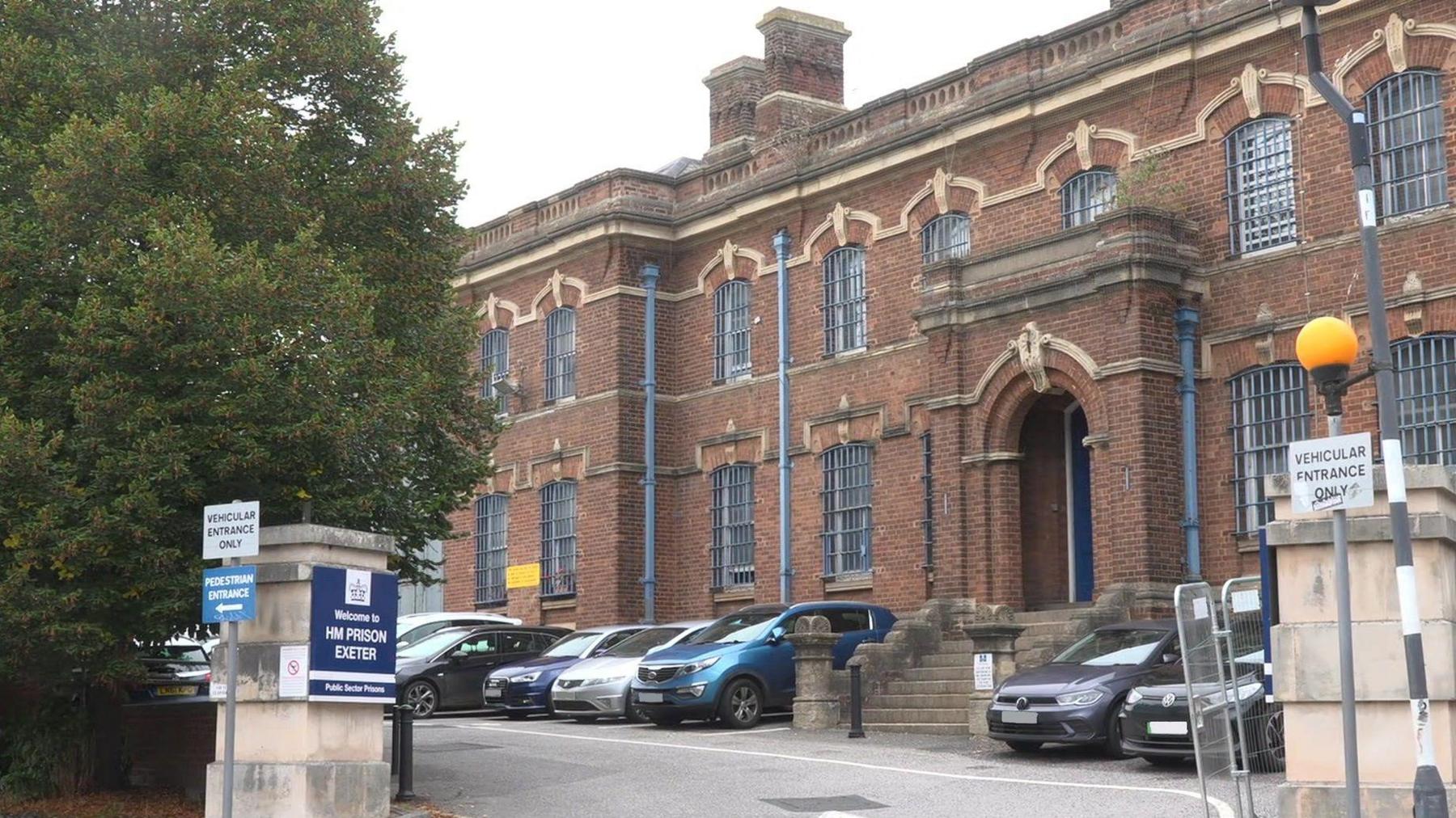 The entrance to and exterior of HMP Exeter on a grey day. On one of the stone entrance pillars is a sign reading: WELCOME TO HM PRISON EXETER and there are two signs reading: VEHICULAR ACCESS ONLY either side of the open entrance. There are cars parked next to the two-storey building which is red stone and looks historic as it has an ornate stone balustrade running around the roof line, many chimneys and pale stone features above the barred windows