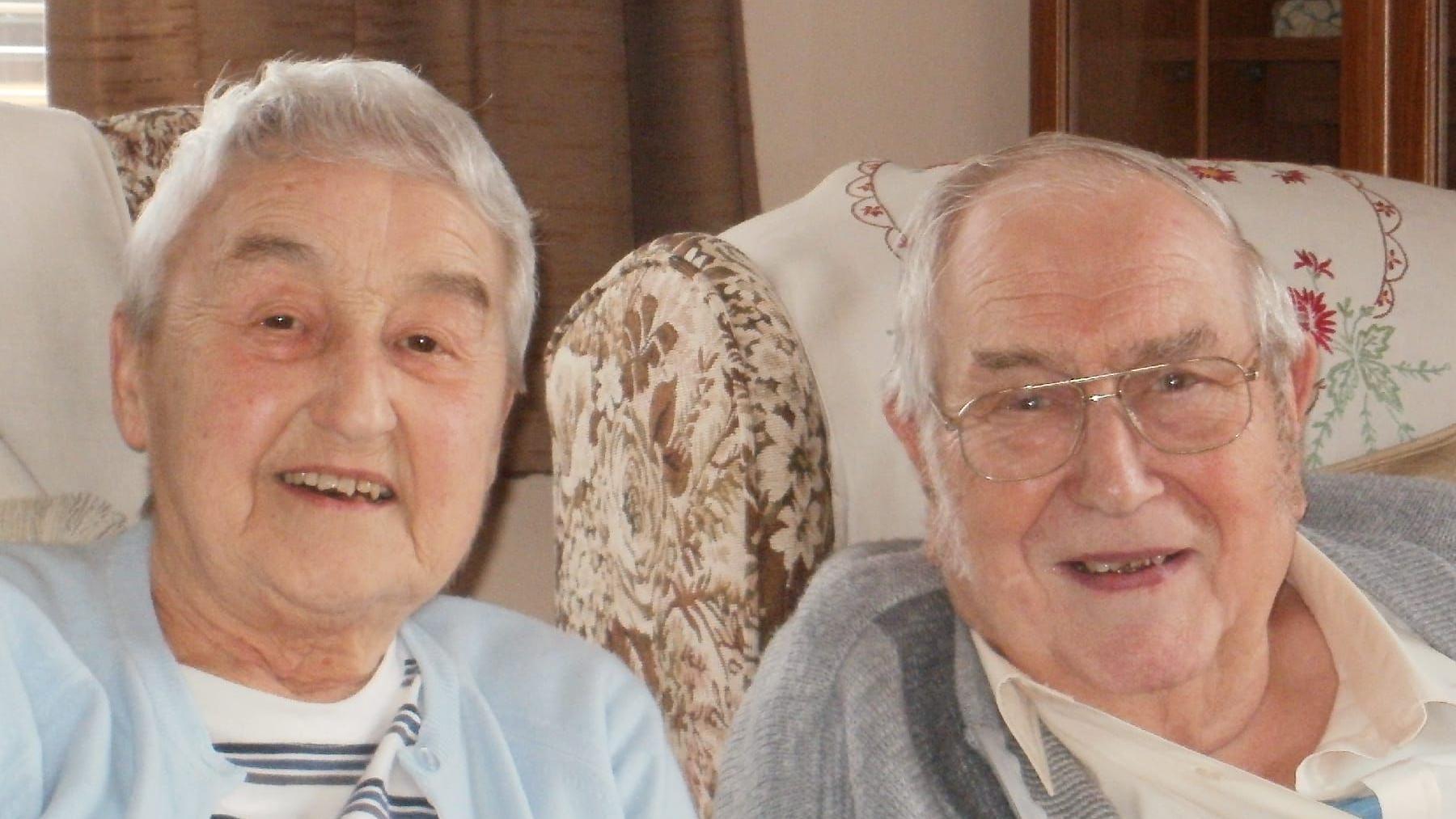 An elderly woman and man smile at the camera as they sit in armchairs. The woman has short grey hair and wears a light blue cardigan and a white top underneath with grey and black stripes on it. The man wears a white shirt with a collar and a grey jumper over the top. 