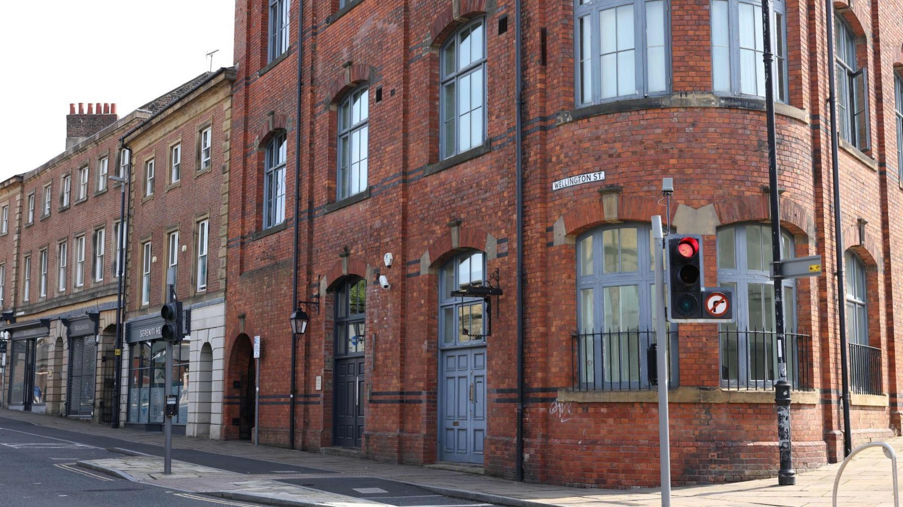 The former Gateshead Railway Club on Wellington Street. It is a large three-storey building made out of dark orange brick with blue-grey window frames. It sits on a corner by some traffic lights, with a curved wall as it bends between the two roads.
