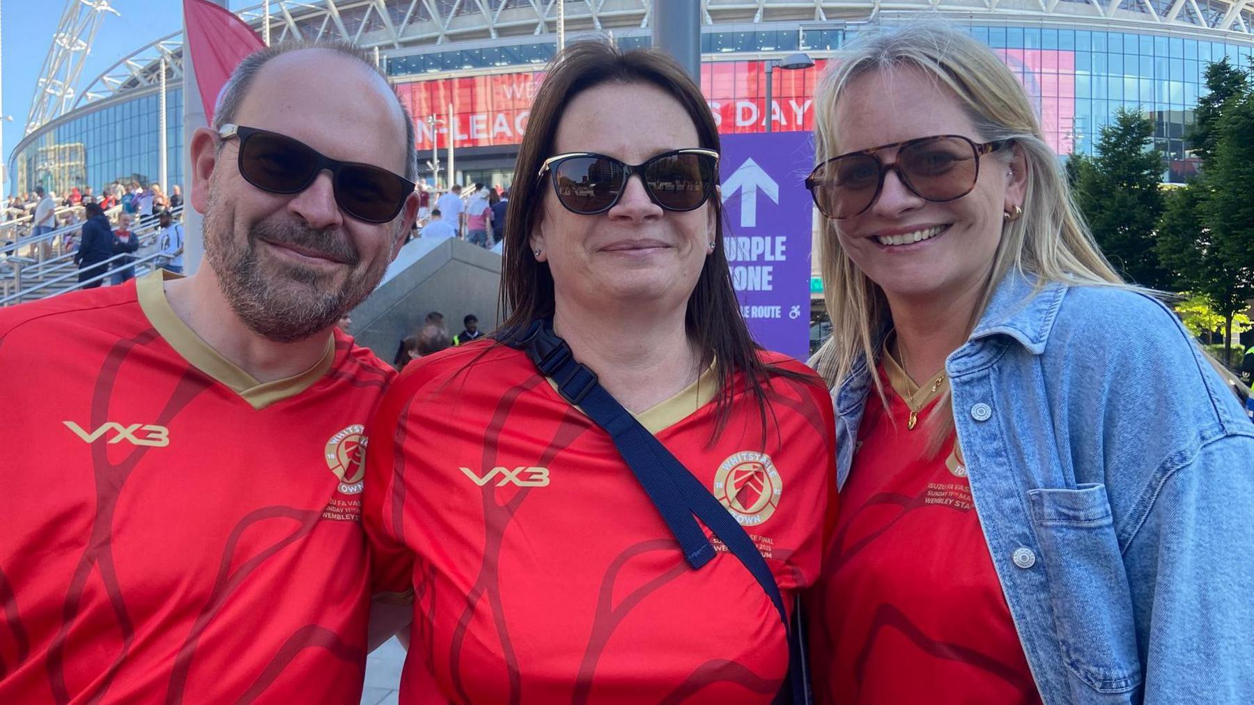 A man and two women wearing red Whitstable Town FC shirts in front of Wembley.