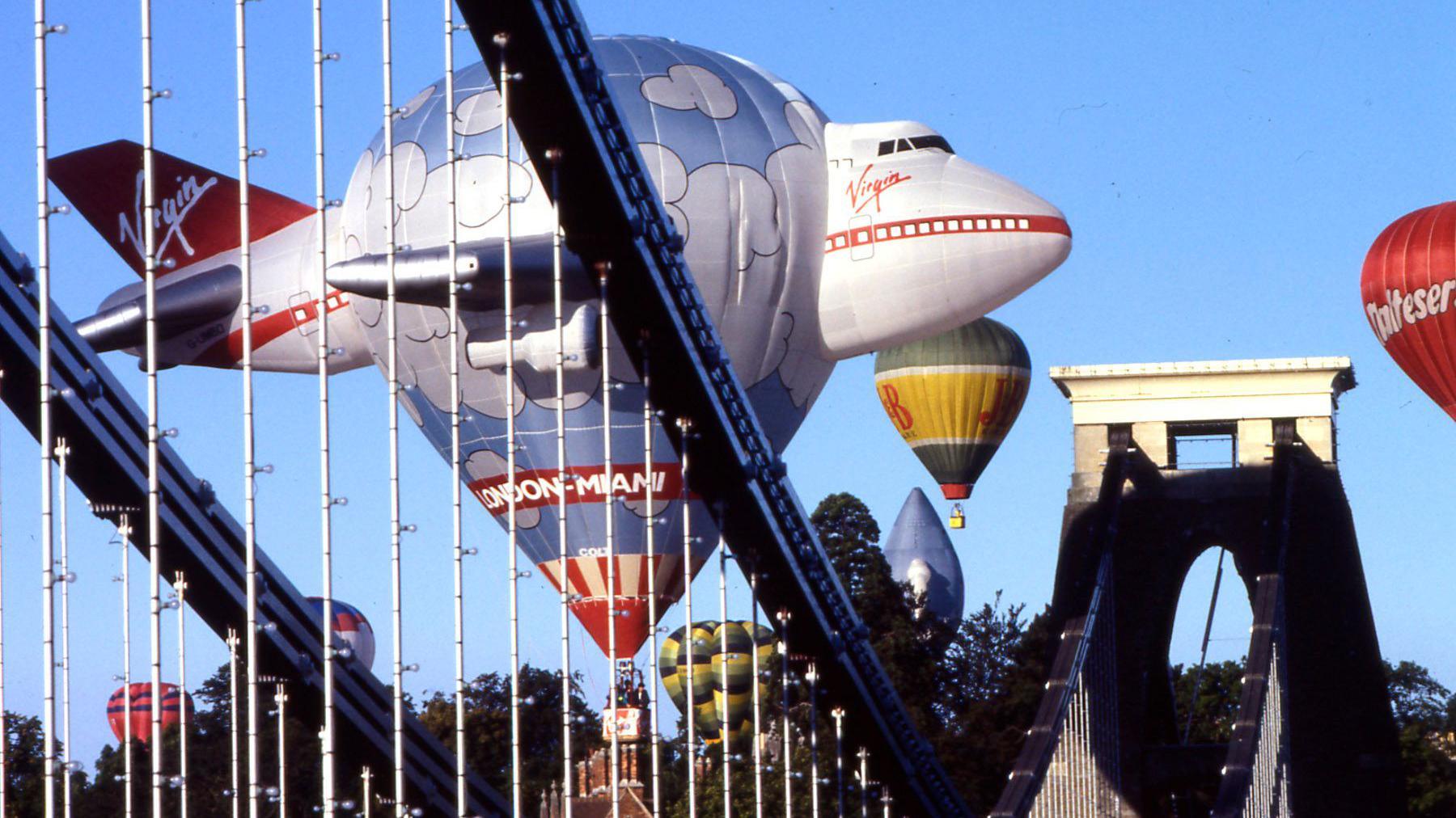 A hot air balloon in the shape of a virgin atlantic airplane flies near the iconic arch of clifton suspension bridge.