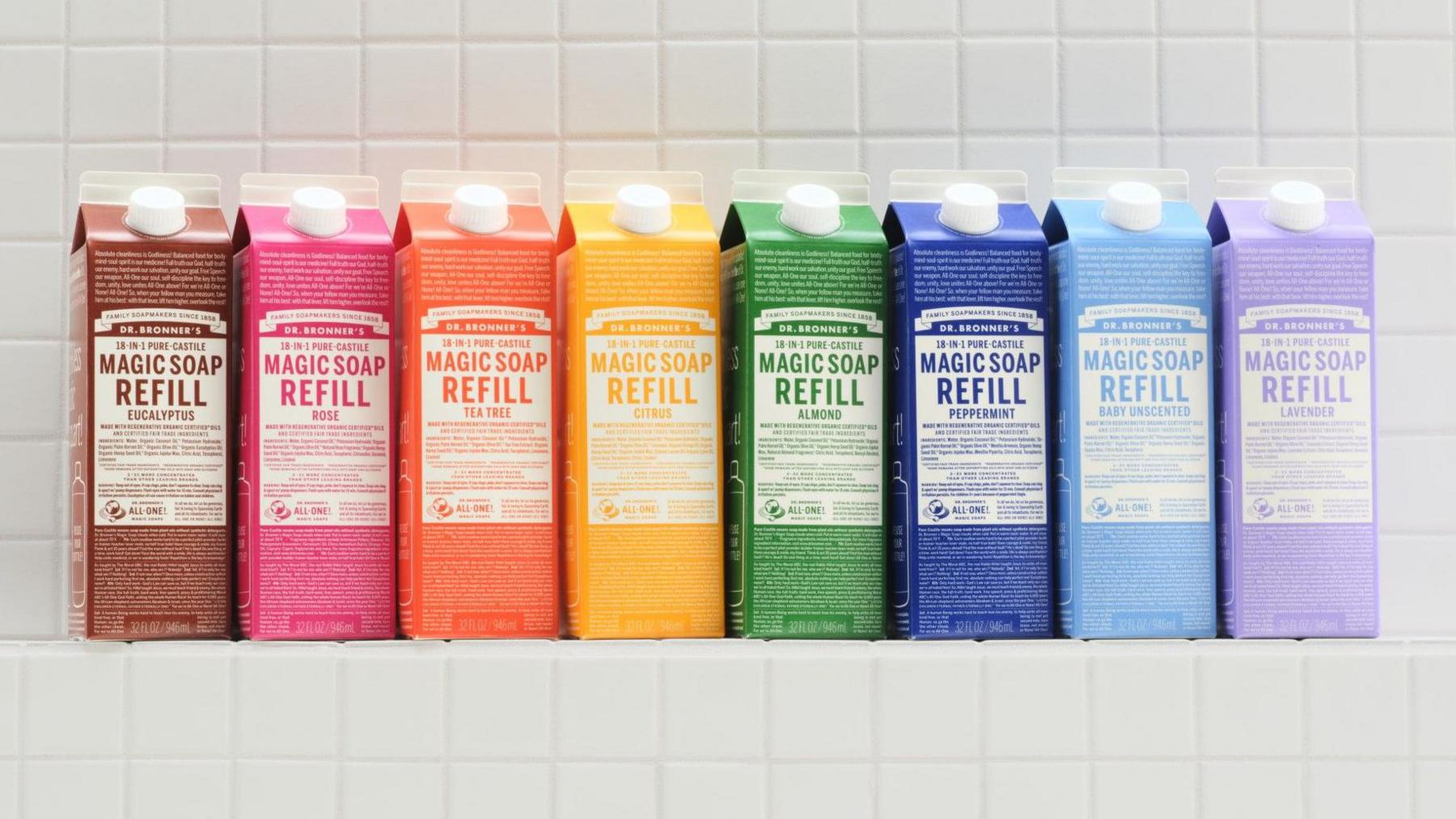 A row of eight refill packs of Dr Bronner's Magic Soap in many bright colours lined up on a white-tiled shelf.