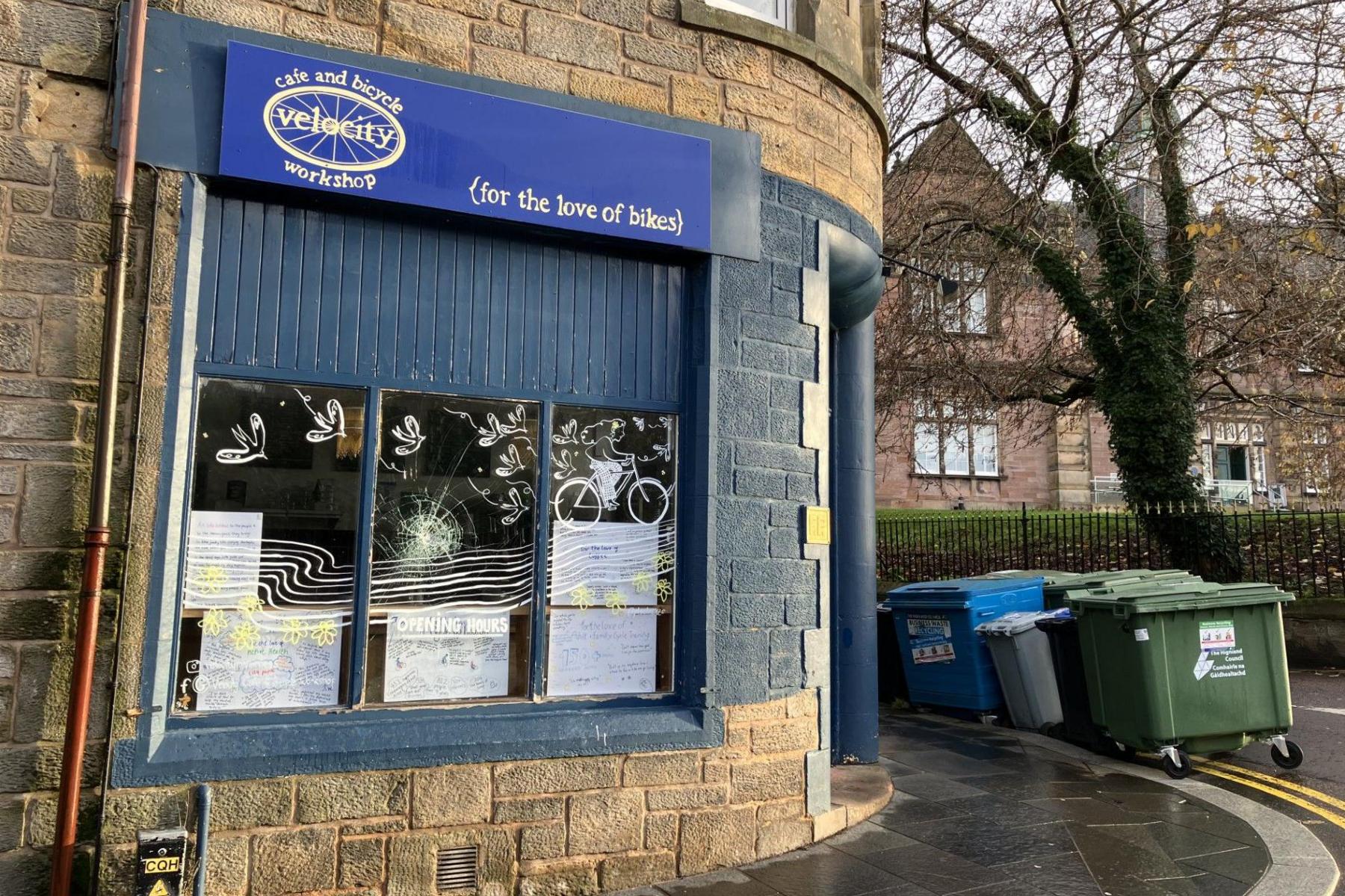 The outside of the cafe and workshop has blue branding. A sign above a window includes the words: "For the love of bikes". By the side of the pavement are industrial size wheelie bins.