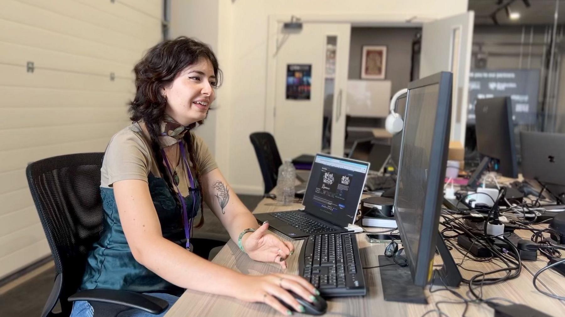 Yasmina Fadel from Maximalist sits at her desk at Production Park in South Kirkby. She is wearing a blue green dress and has a leaf tattoo on her left arm. She's got the computer mouse in her right hand.