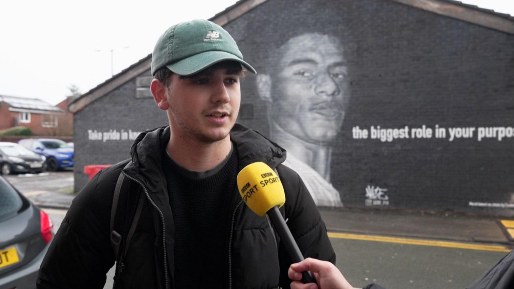 Manchester United fan Brandon Gittins stands talking to a BBC reporter in front of a Marcus Rashford mural