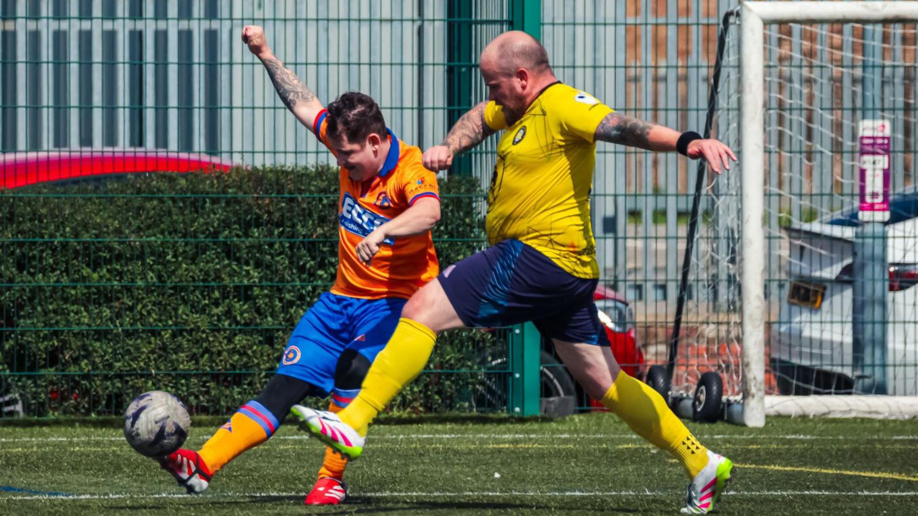 A clash between two footballers, one in an orange Teesside Erimus kit and the other in yellow. The Teesside player is mid-kick and the player in yellow is lunging towards him to intercept the pass. A goal can be seen behind them to the right.