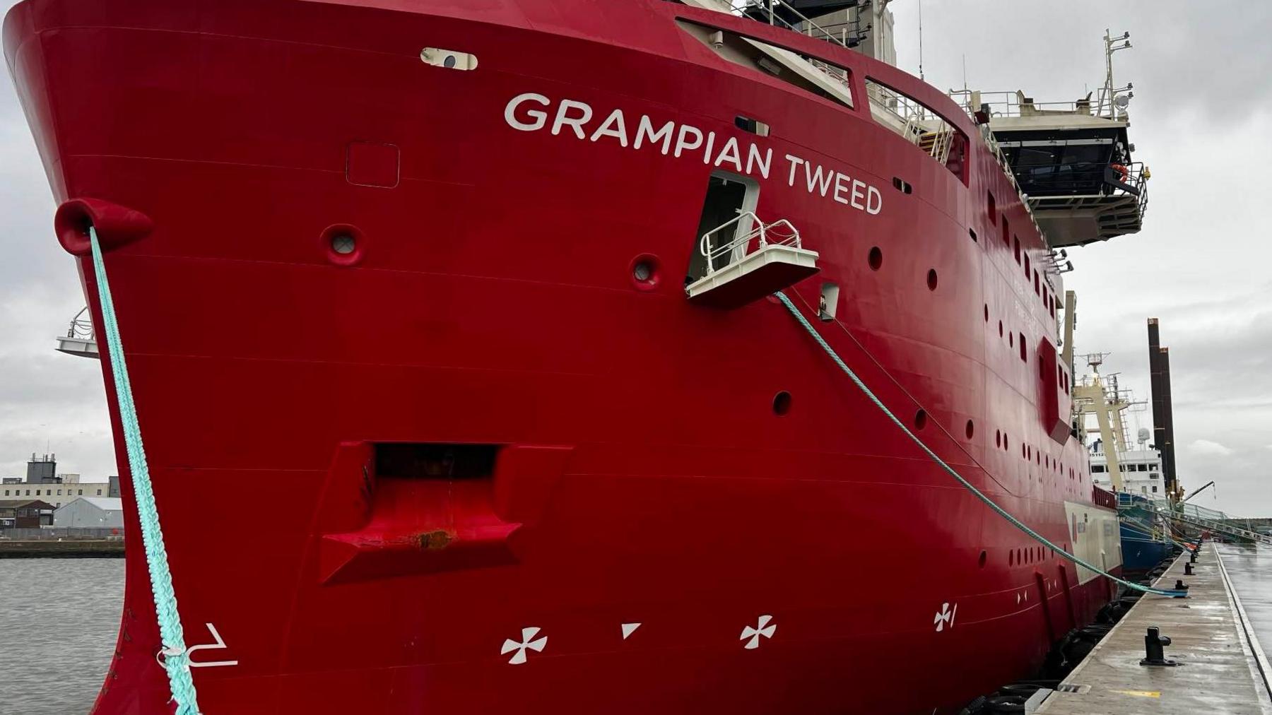 Grampian Tweed is a large red ship seen docked at a port. A white gang plank is open halfway down the side of the ship. 