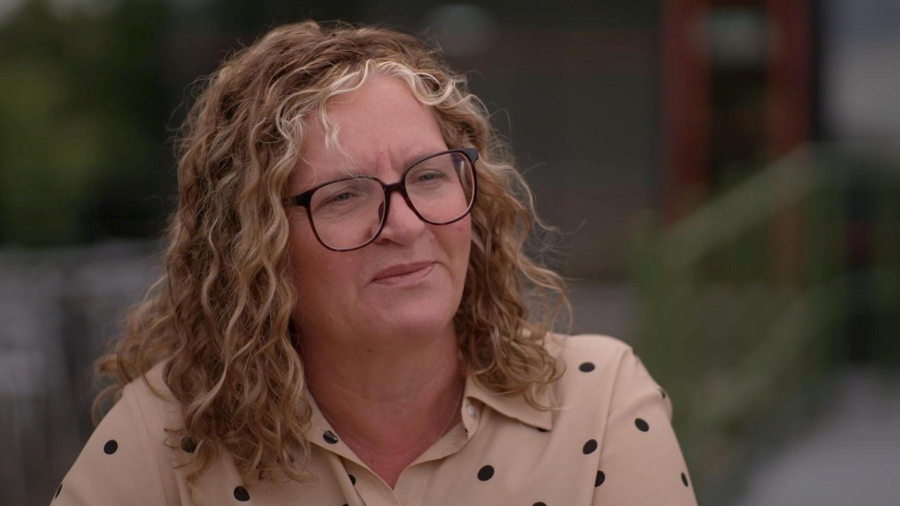 Catherine Marchant has enarthrosis magnitude blonde wavy hairsbreadth and achromatic rimmed glassed. She wears a beige garment with achromatic polka dots. She looks heavy successful thought, distant from the camera.