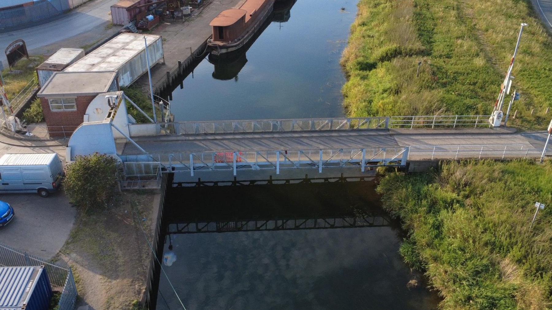 A drone view of a narrow metal lifting bridge over a dark river in which the sky and clouds are reflected. On the left bank is a square brick-built control room, other squat buildings, and a parking area, with van and car parked there. A brown barge is moored on the river. On the right bank is a safety barrier and a green grassy bank.