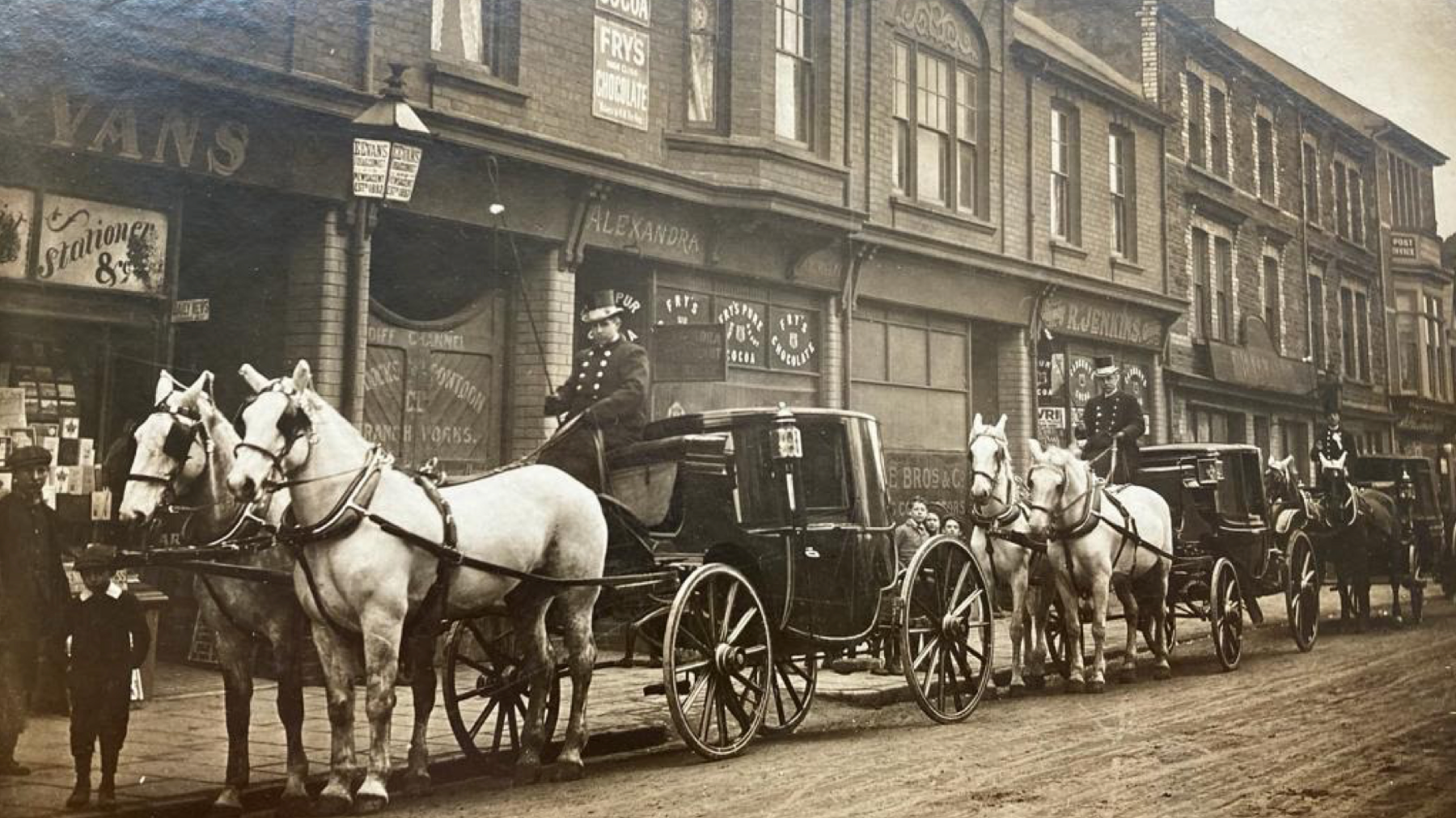 Pictured here is the wedding procession of Max and Mabel Wulff. Six large black and white horses pull three carriages along Alexandra Road in Pill, South Wales. The photograph is black and white and depicts a typical high street in the UK at the turn of the century.