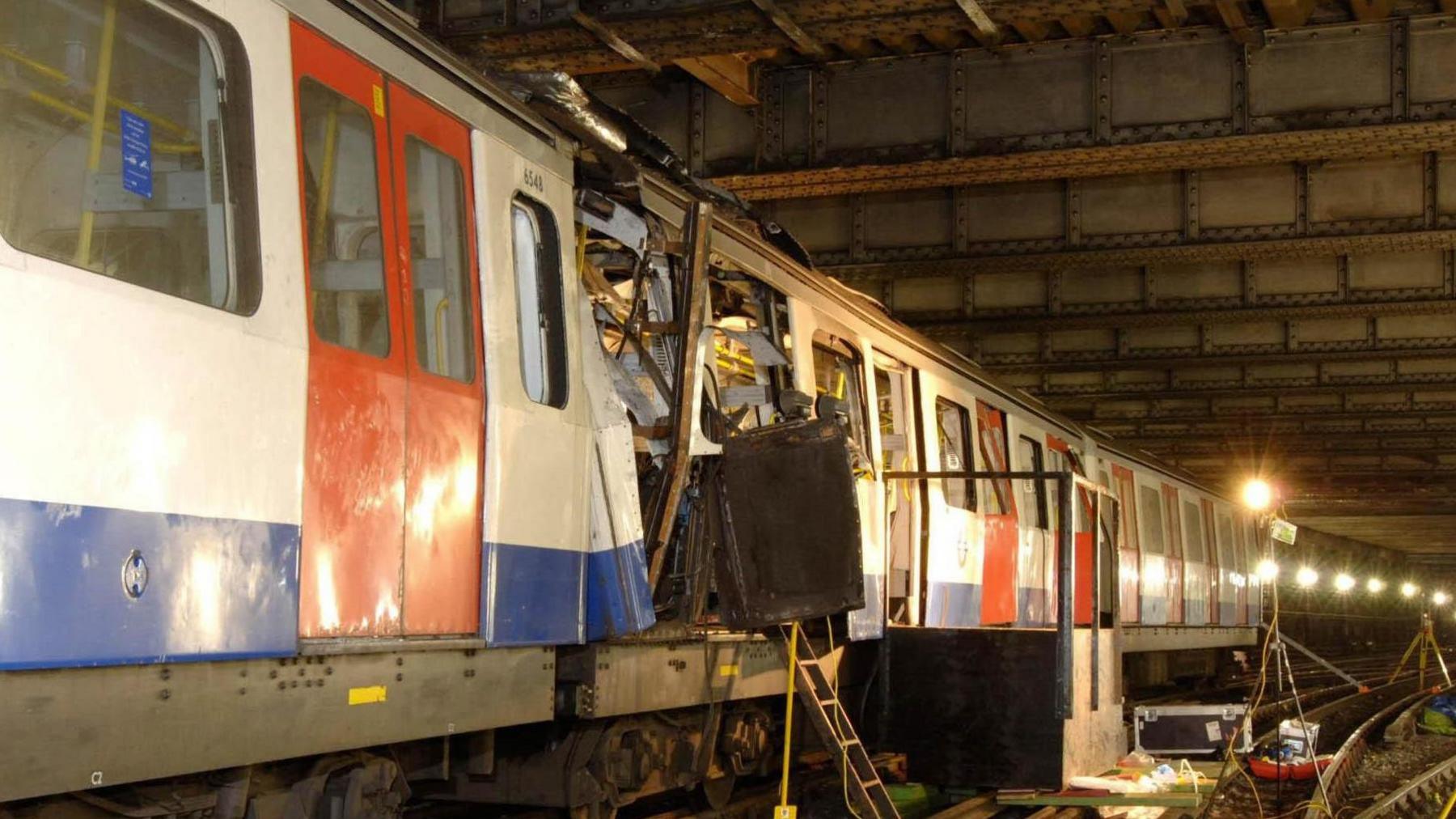 A blue and white London underground tube train with red doors is pictured underground. Part of the carriages are damaged in the middle of the train with a door and metal hanging out. A light and a ladder can be seen pointing into this section of the train with other equipment on the train tracks.