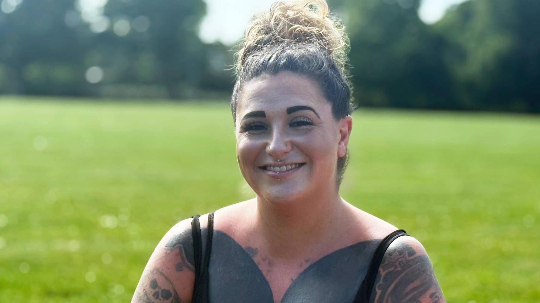 A smiling woman with heavy tattoos sits on some grass.