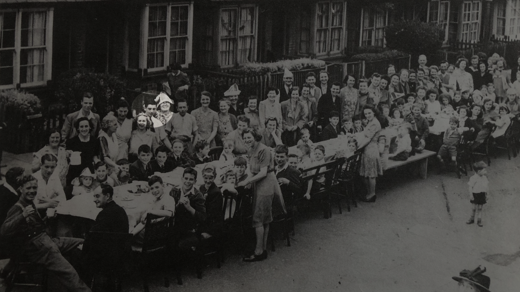 A black and white wide shot of a VE Day street party. Barry is standing wearing a white shirt and black tie, smiling into the camera.