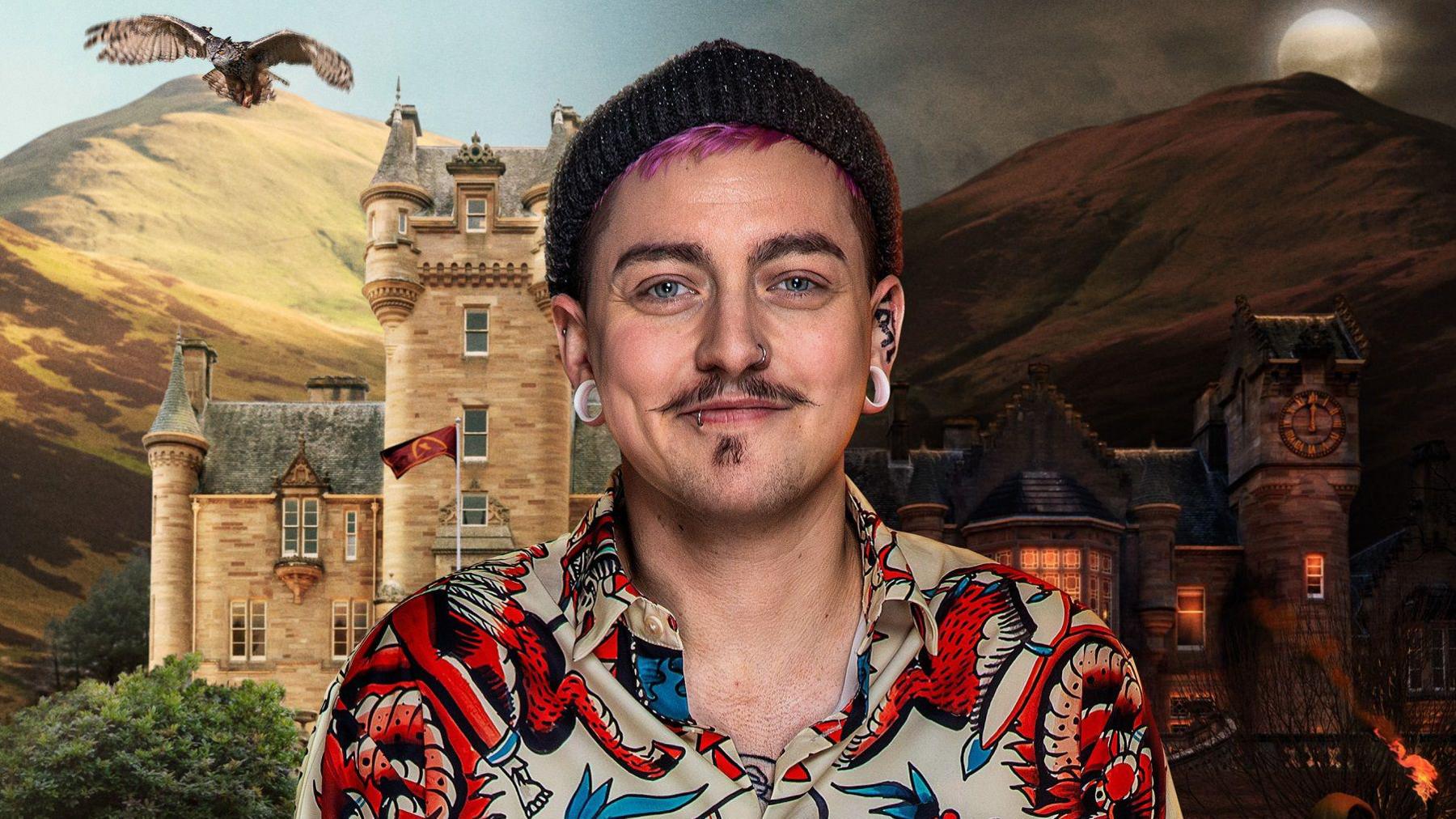 A man with pink hair and a dark moustache wearing a beanie hat and a colourful shirt. Behind him a background shows a castle in the Scottish highlands.