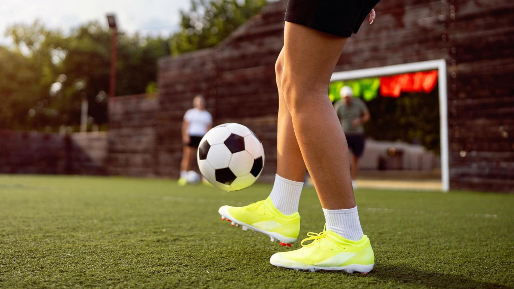 An unrecognisable teenage female football players juggling the ball during training on a green pitch.
