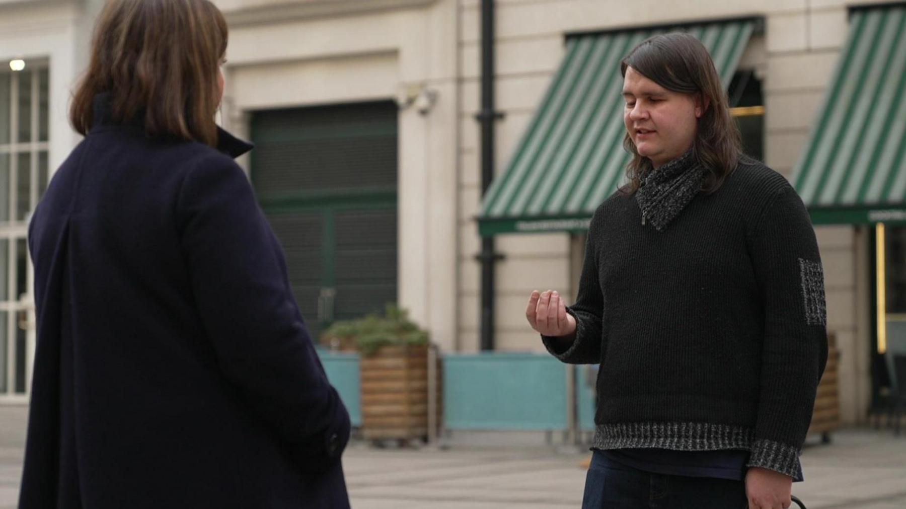 Kelsey is wearing a black knitted jumper with dark grey trims. They have long dark hair and they are talking to a reporter who is interviewing her, with her back to the camera. 