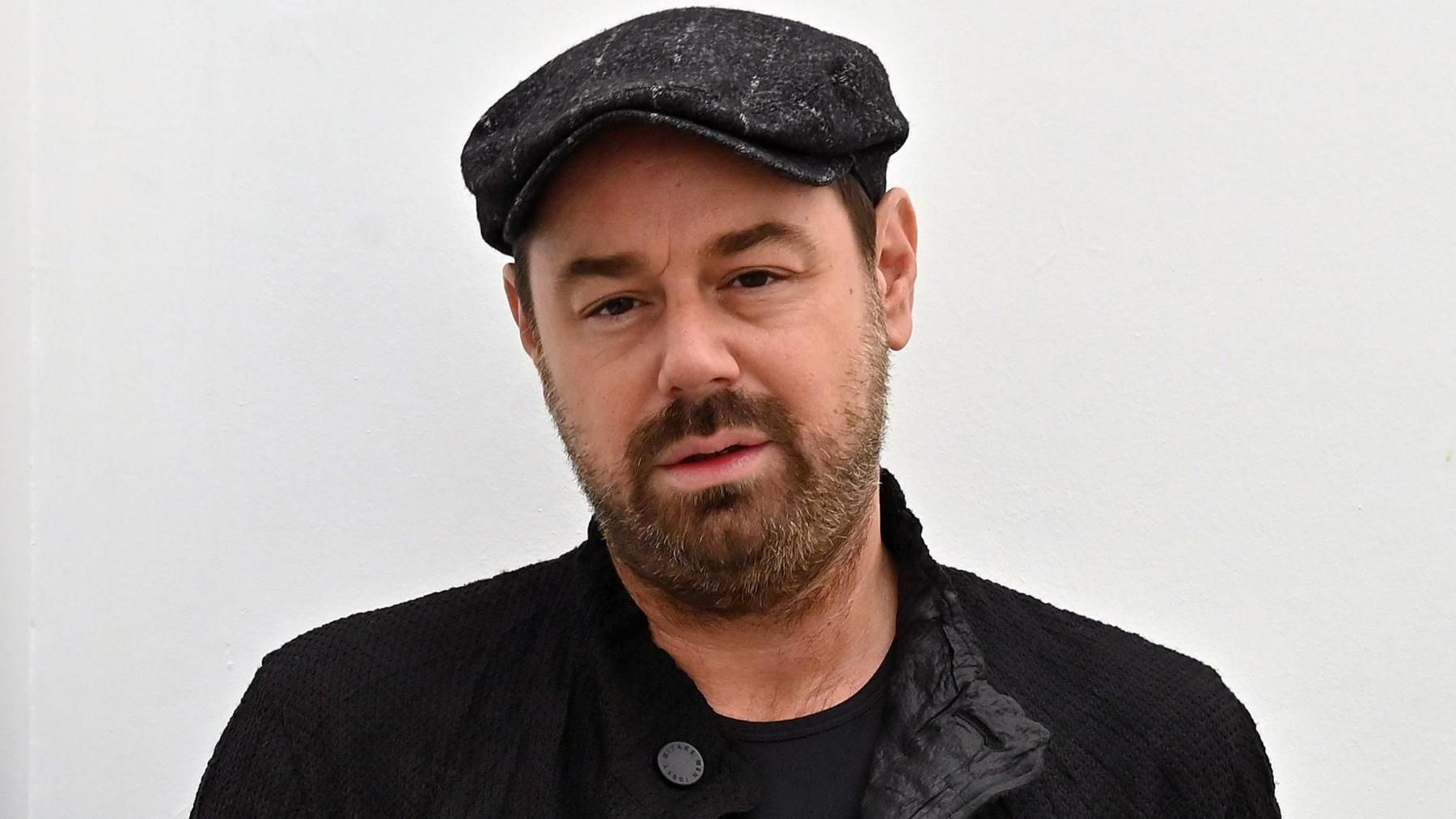 Danny Dyer stood in front of a white wall. He is wearing a black top, jacket and hat.