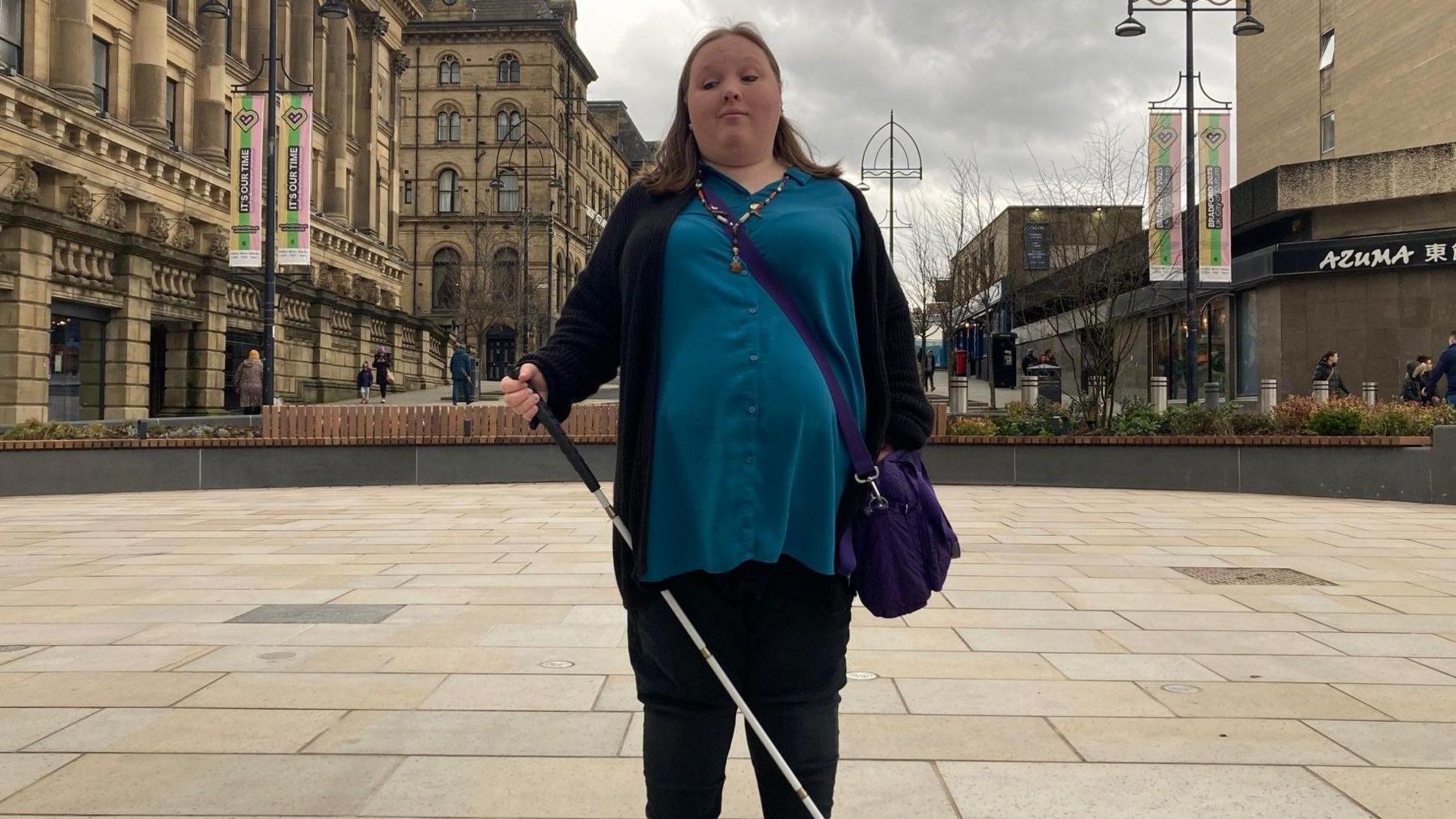 A woman with a white stick standing in a paved area with buildings on either side of her.