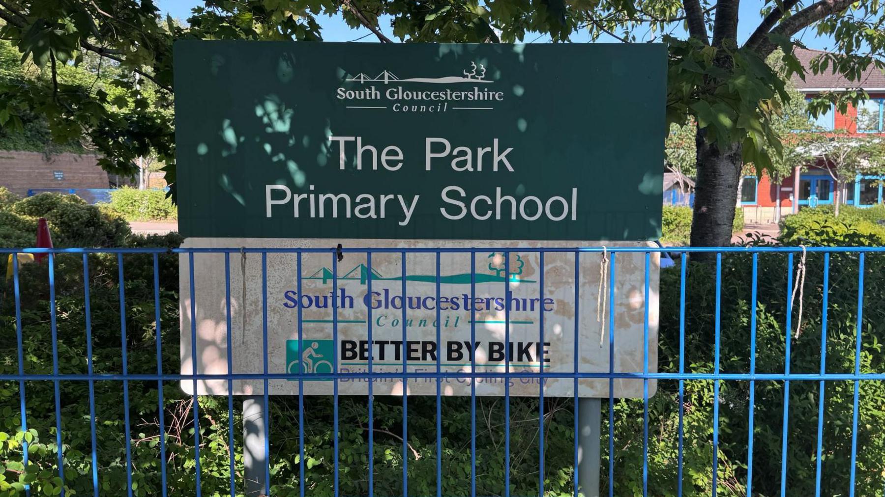A sign for the Park Primary School, with white letters on a green background, behind a thin blue metal railing fence