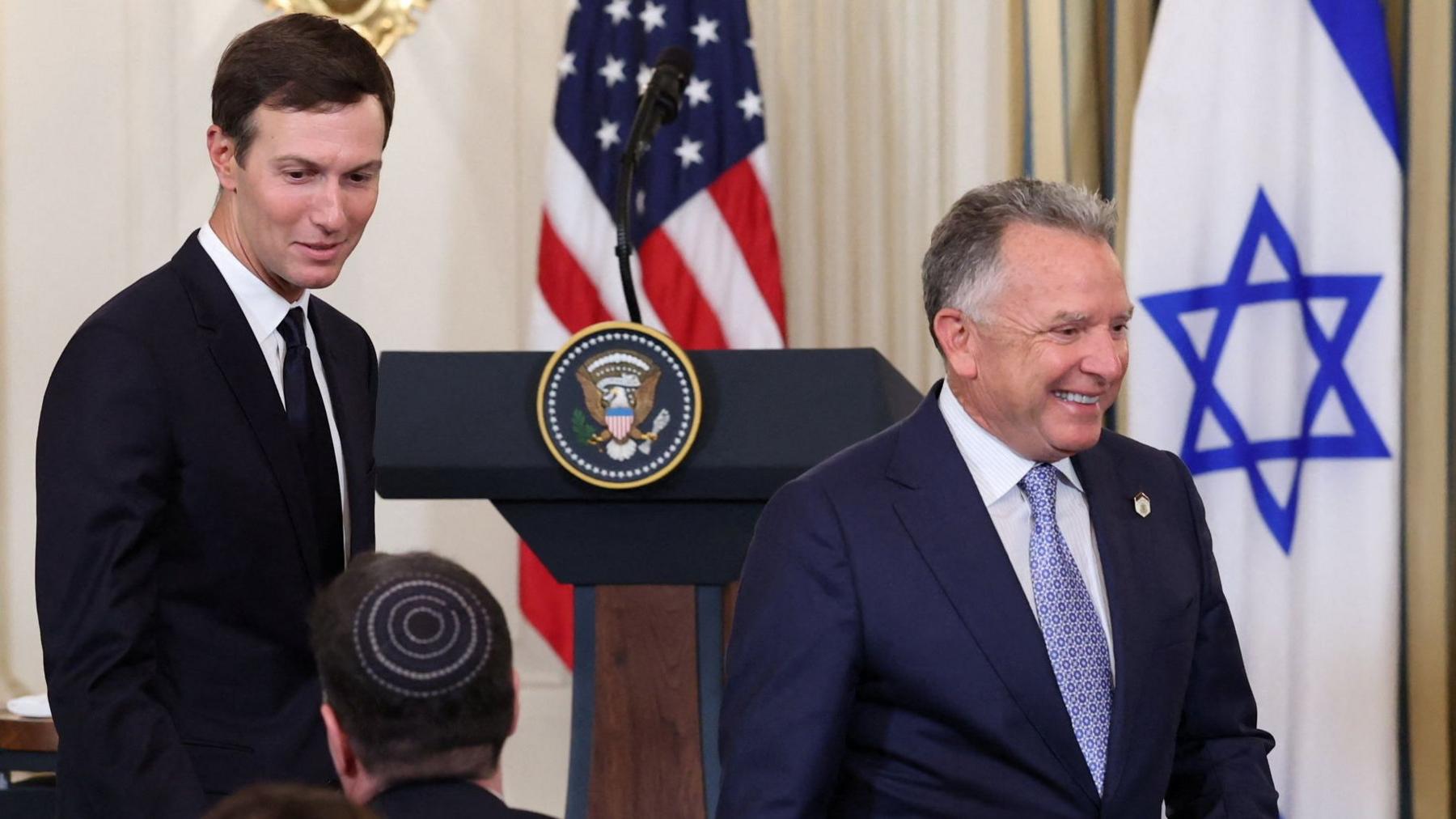 Jared Kushner, wearing a dark suit and tie, and Steve Witkoff, wearing a navy blue suit and light blue tie, walk through a press conference. A US and Israeli flag are in the background.