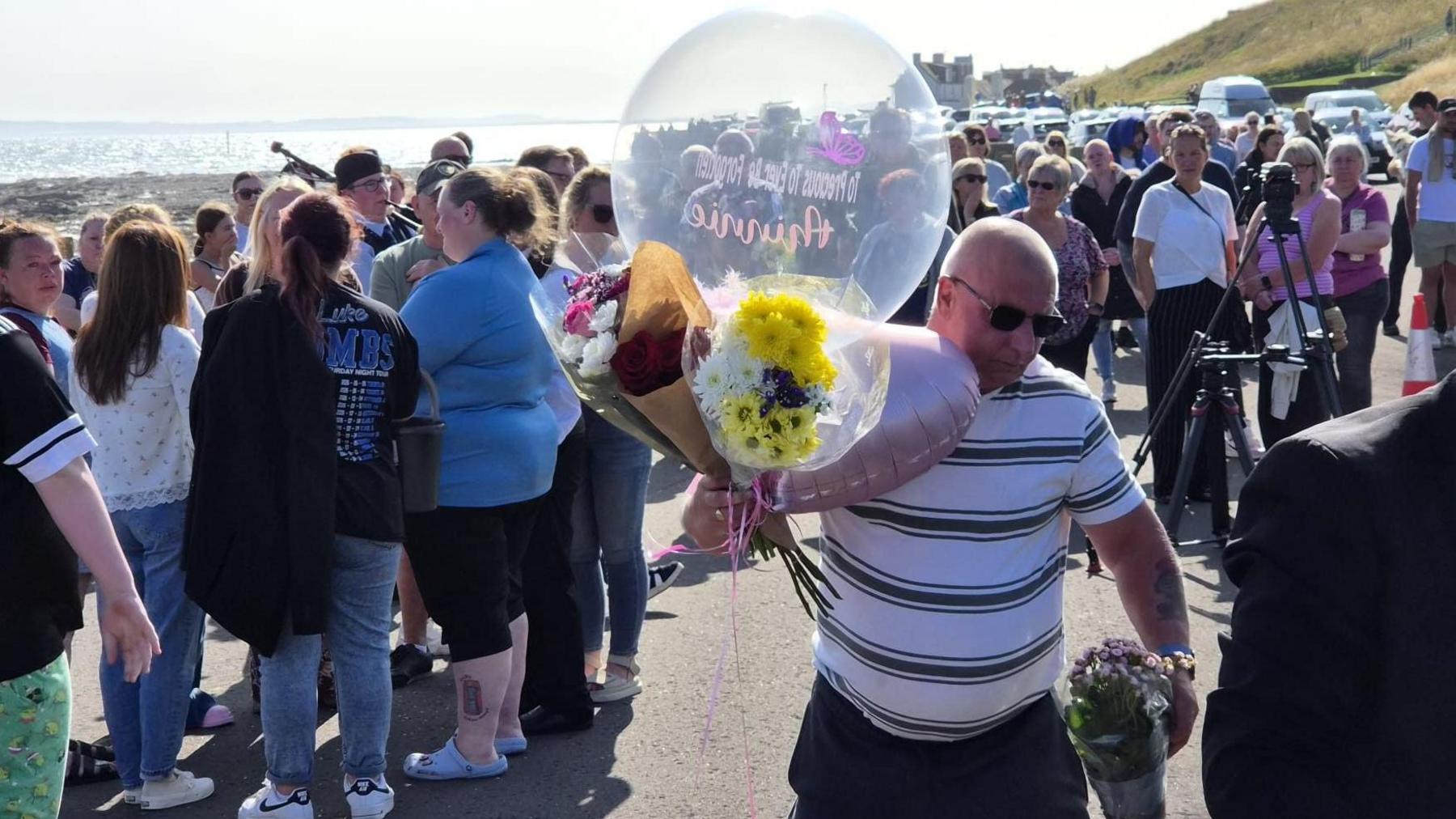A crowd of people at a beach. A man with sunglasses holds a balloon and a bunch of flowers