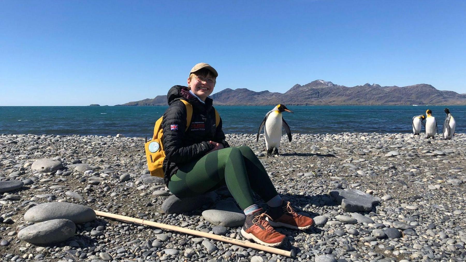 Aoife McKenna in Antarctica. She is wearing a cap and glasses, and sitting down on a rock by the shore, with four penguins and the ocean behind her 