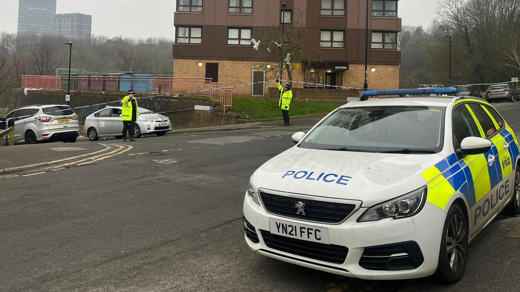 A police vehicle is parked on the left  with a cordon extended around the junction. Two officers can be seen standing guard.