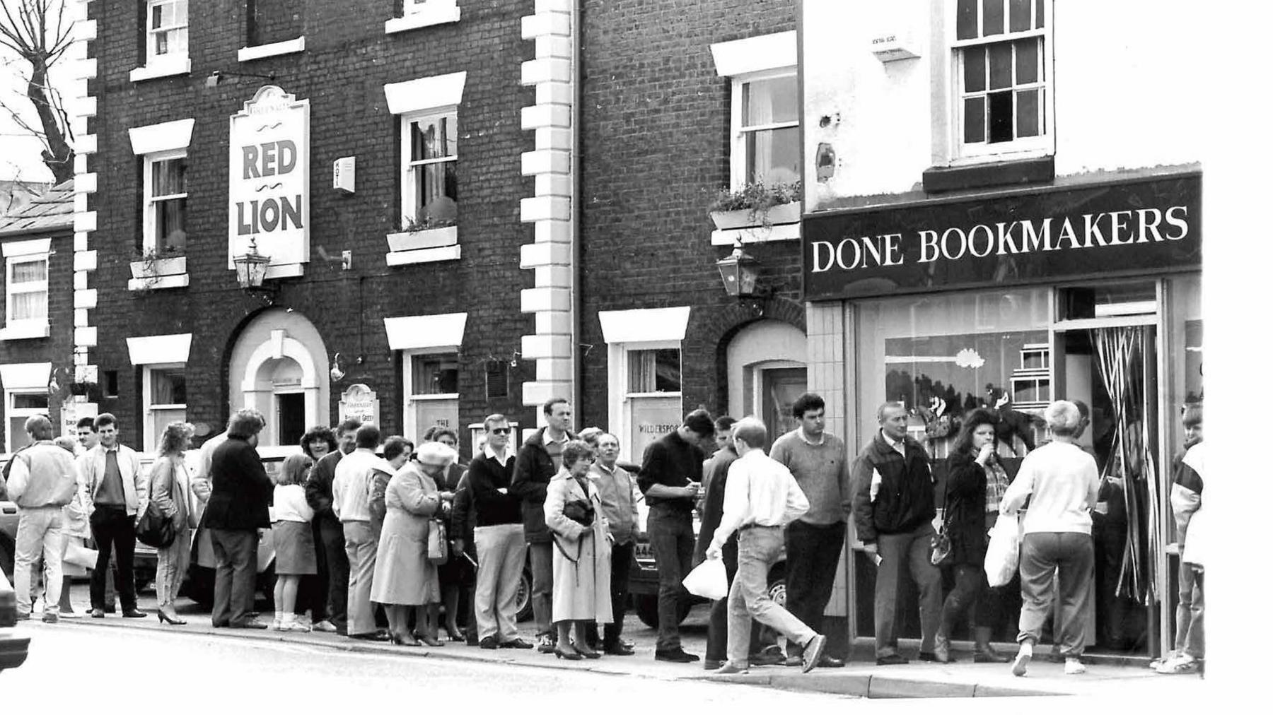 A black and white photograph of men and women, wearing casual clothes, queuing on the street outside a Done Bookmakers shop and in front of the neighbouring Red Lion pub