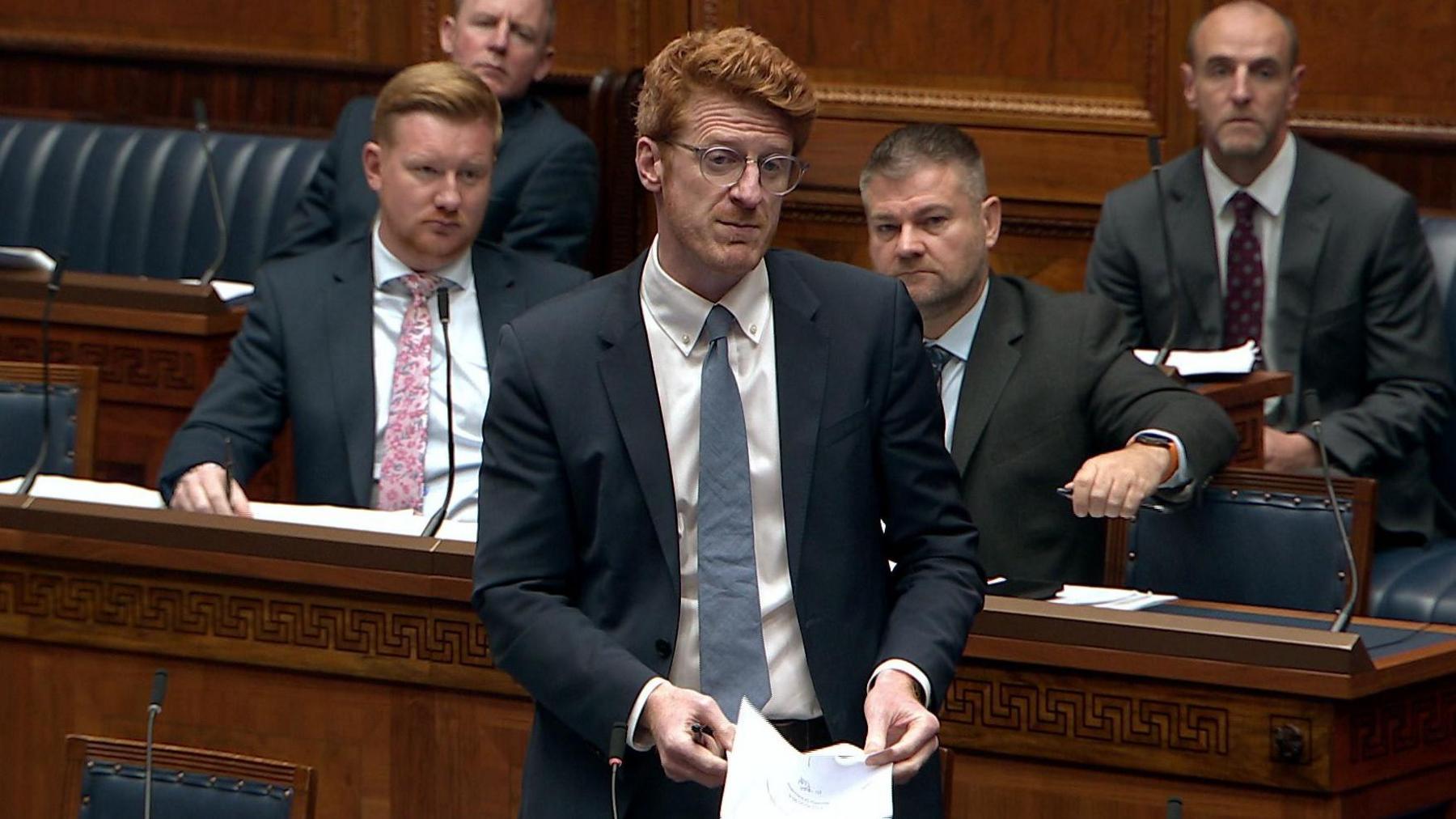 Matthew O'Toole, a man with longer, ginger hair, is standing in the Stormont chamber making a speech. He is wearing a pair of circular glasses, a navy suit jacket, a white collared shirt and a light blue tie. He is holding some sheets of paper. Behind him are fellow party members of the SDLP - who sitting behind the benches of the chamber on blue, leather seats.