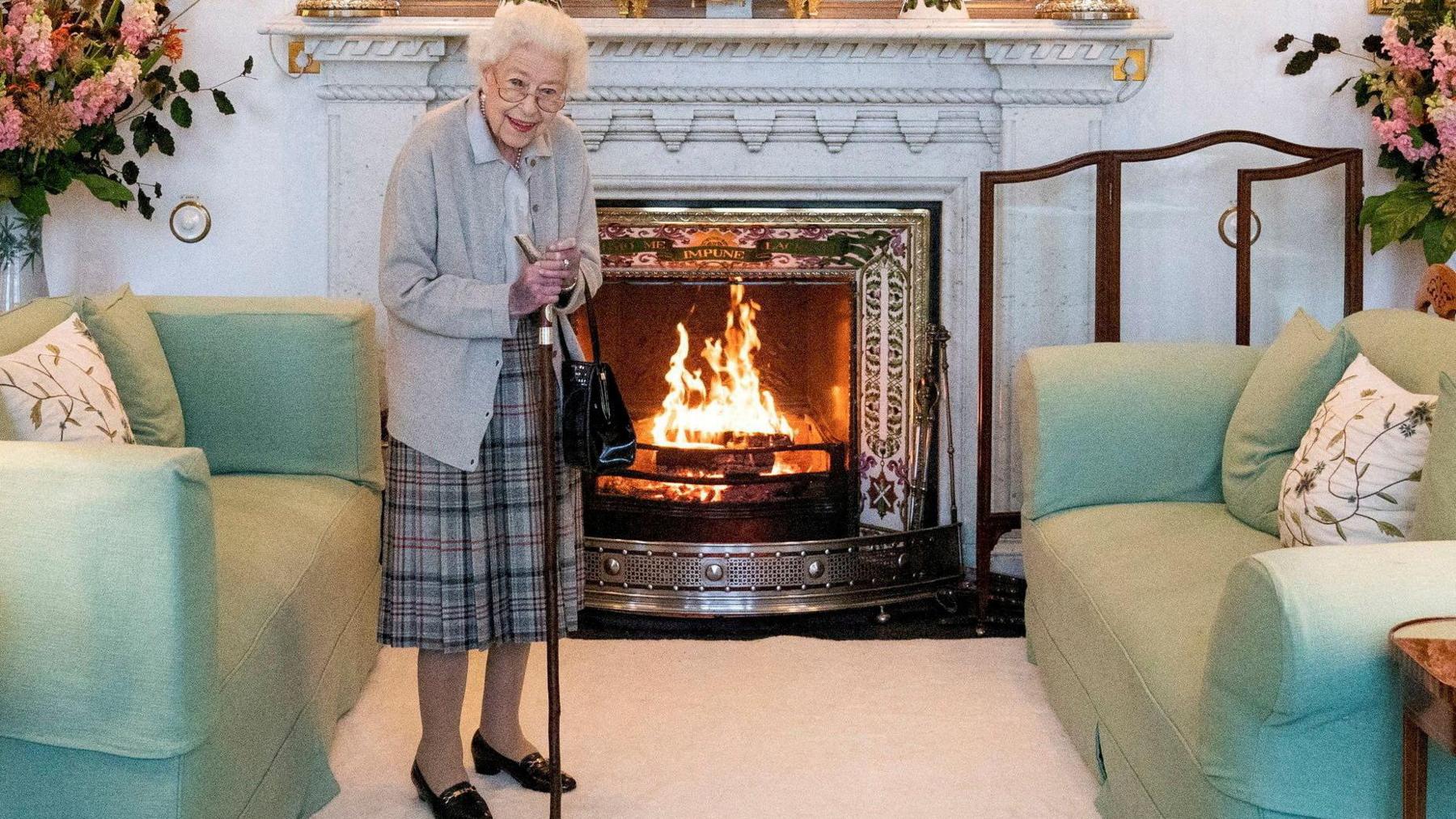 The late Queen Elizabeth stands in front of a fireplace at Balmoral. She has short white hair and wears glasses. She has a walking stick and wears a grey cardigan and a tartan pleated skirt