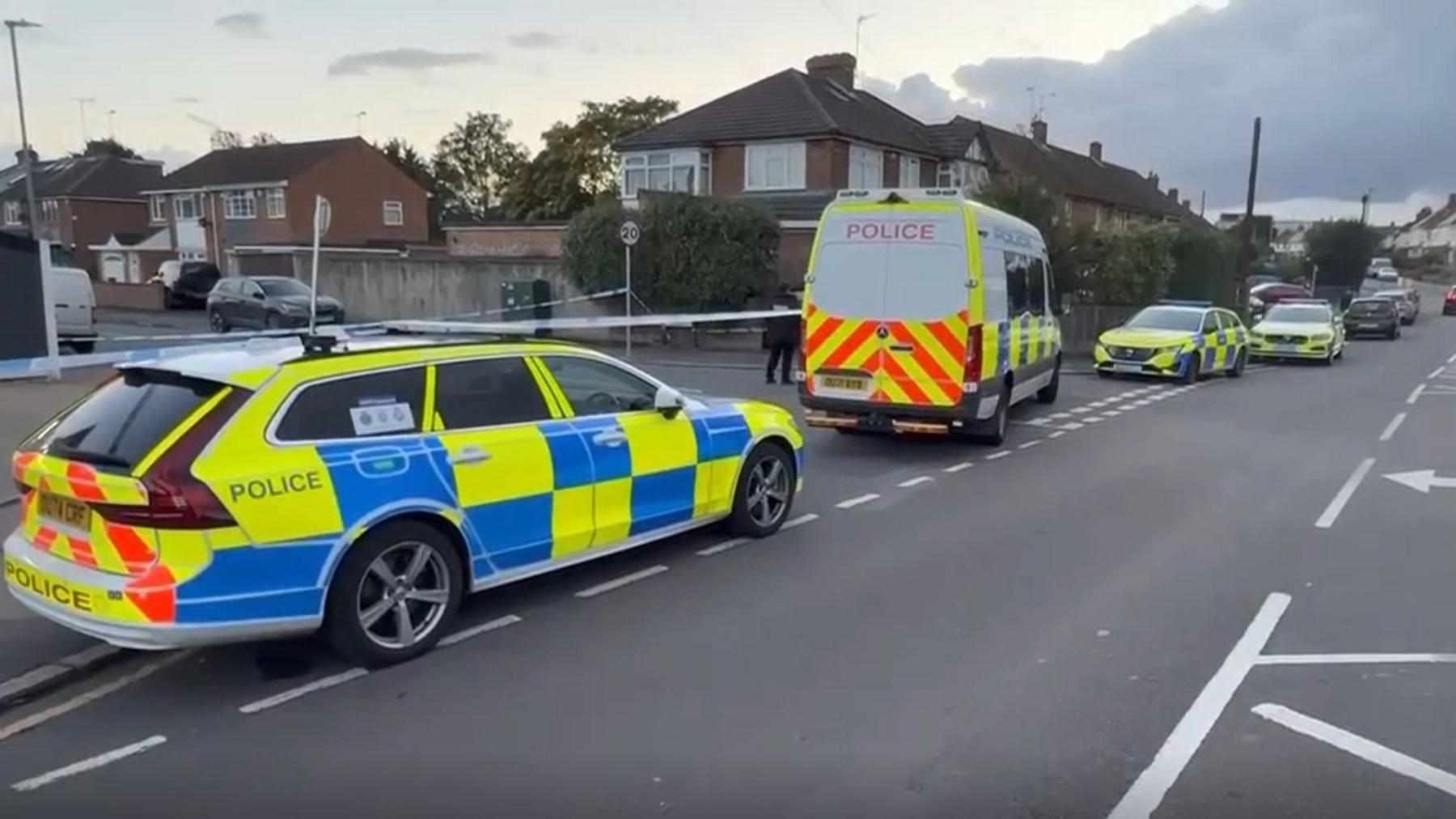 A residential street with multiple police vehicles, including three marked cars and a van, parked in a line. Blue and white police tape cordons off entry to a side road.