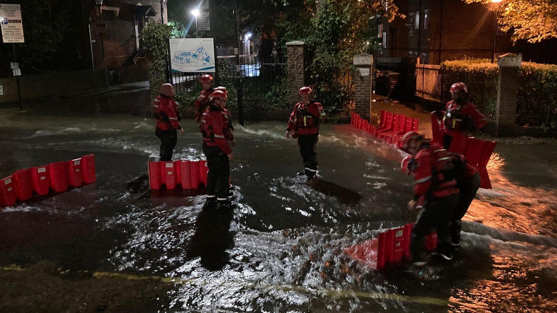 A group of people in high viz clothes standing in a flooded road erecting red plastic barriers
