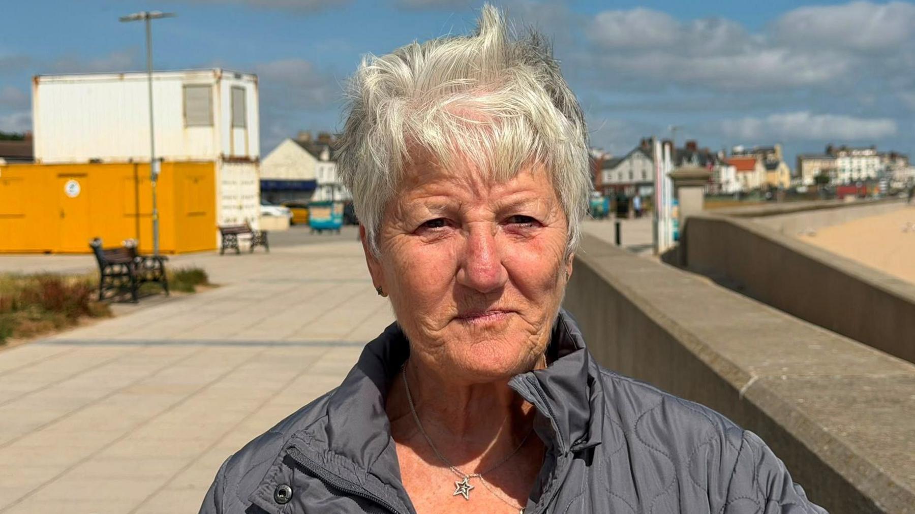 Sandra Gleeson has short white hair. She is wearing a black puffer jacket and a silver necklace with a star pendant. 