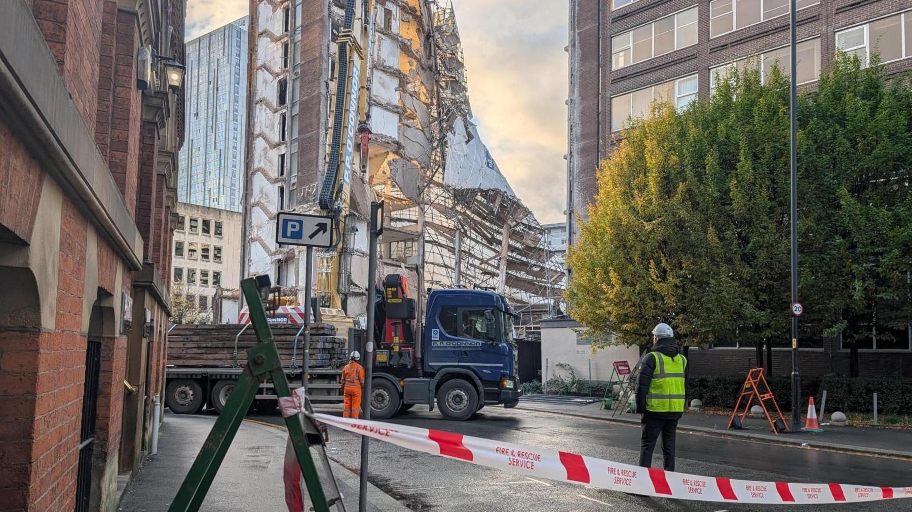 Police tape cordons off the approach to a partially collapsed building. A truck carrying scaffolding can be seen on the road nearby.