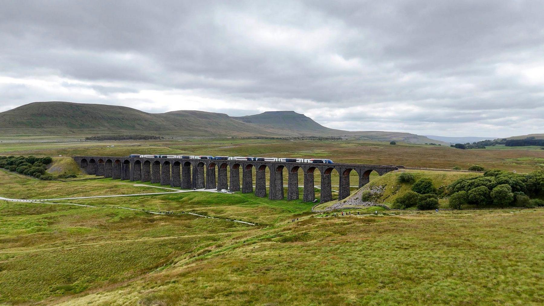 A zoomed out view of the train travelling along the viaduct. Three large hills can be seen in the background. The sky is gloomy and covered in white/grey clouds.