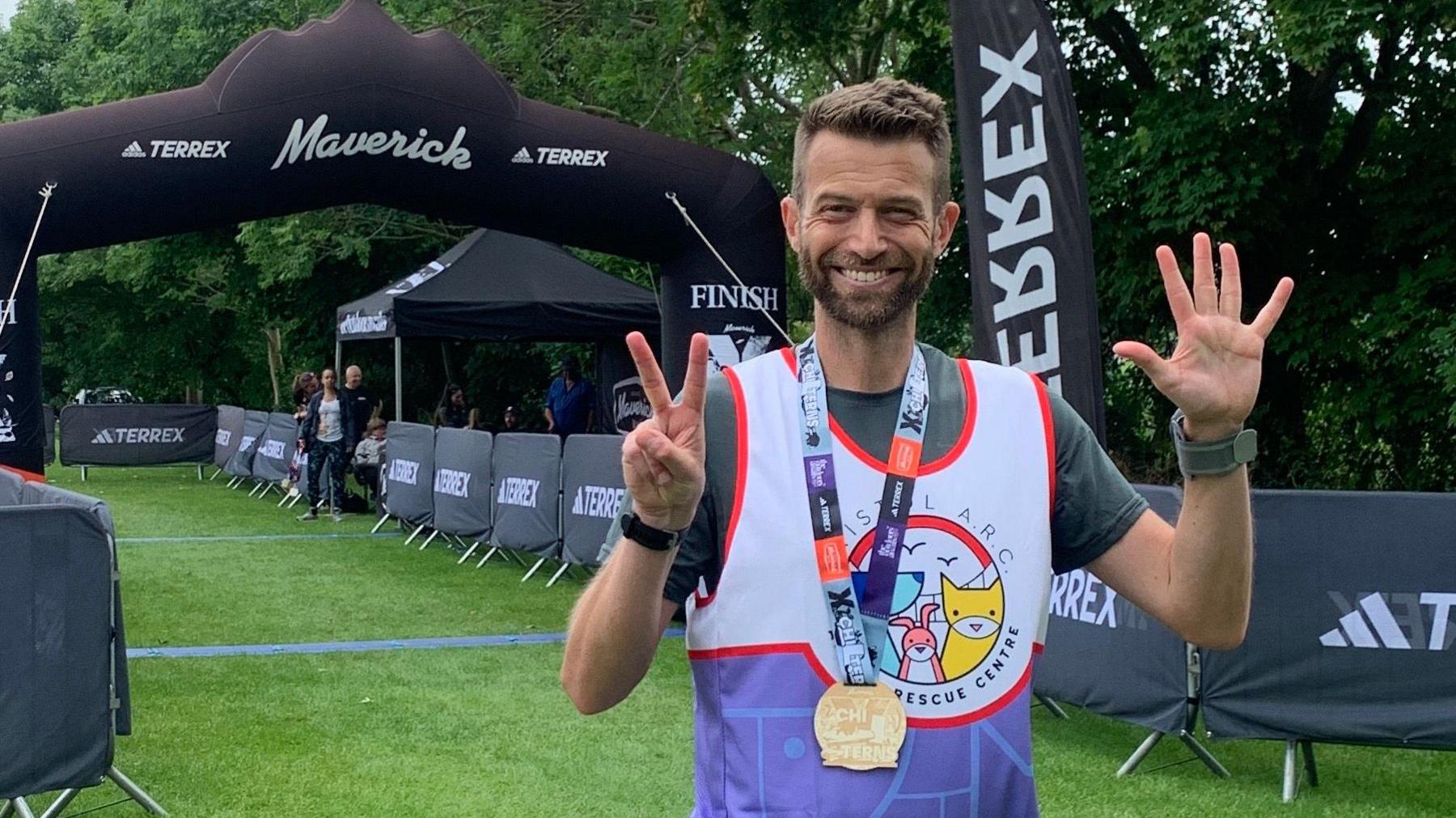 Phil Hobbins-White wearing a Bristol Animal Rescue Centre running vest with the logo on the front. He is wearing a medal around his neck and holding up seven fingers to symbolise it's his seventh marathon this year. He is standing at the finish line of the Maverick Chilterns race.