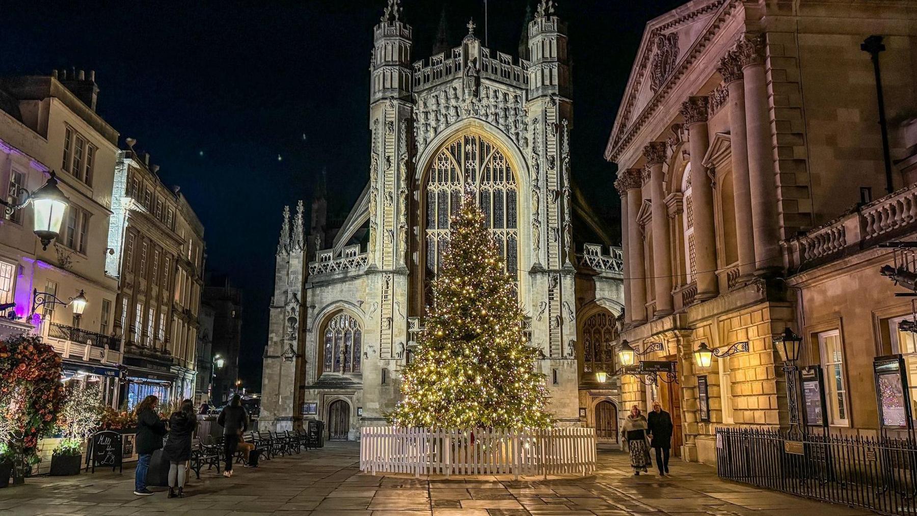 A large illuminated Christmas tree is seen in front of Bath Abbey in the city centre. Small groups of people are looking at the tree. The photograph is taken at night with lights from local businesses glowing and the abbey also lit up