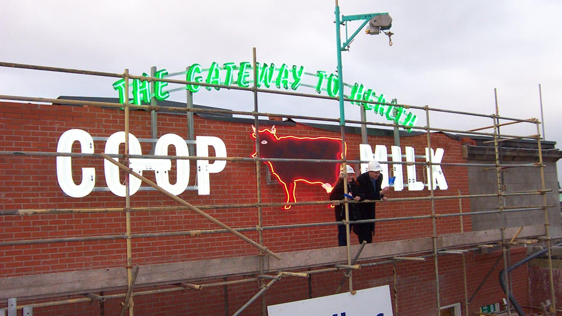 A sign with green lettering and lights saying "The Gateway to Health" and white lettering and lights saying "Co-op Milk". In the middle is a cow figure, which is lit up with red neon. It is on the side of a brick building 