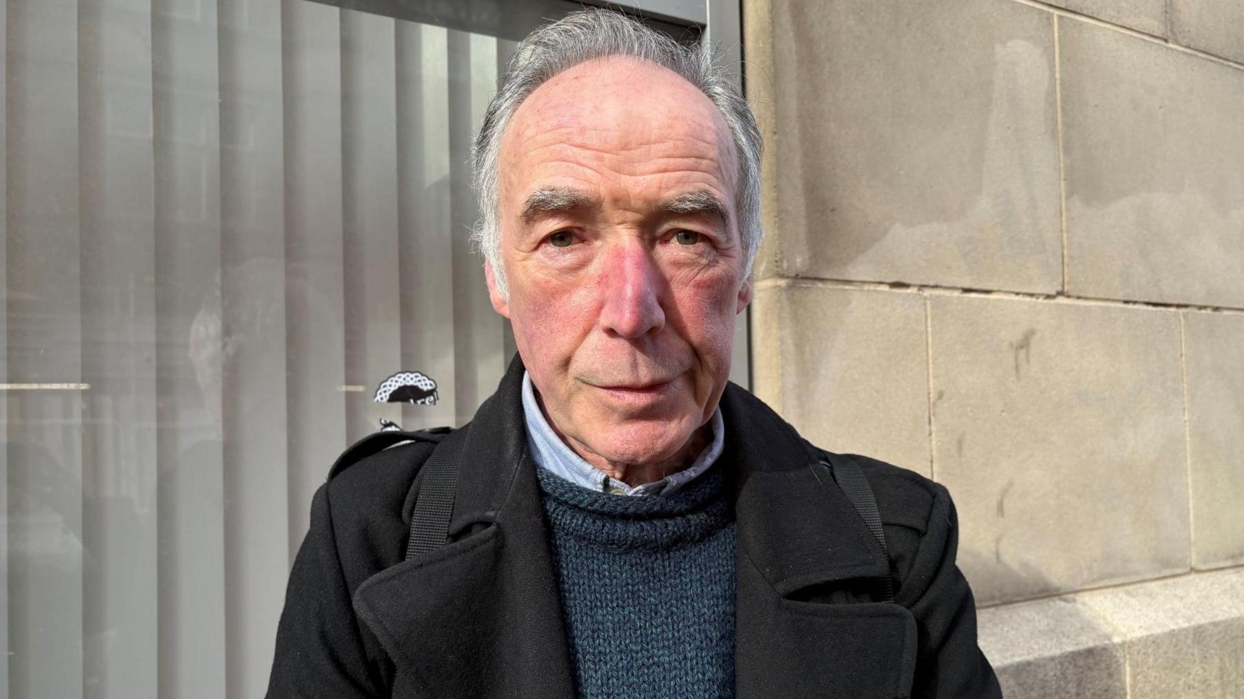 Ciaran Molloy - a man with short grey hair looks at the camera. He is wearing a black coat, a blue jumper and a blue shirt underneath. He is standing in front of a large window and a white-stone building.