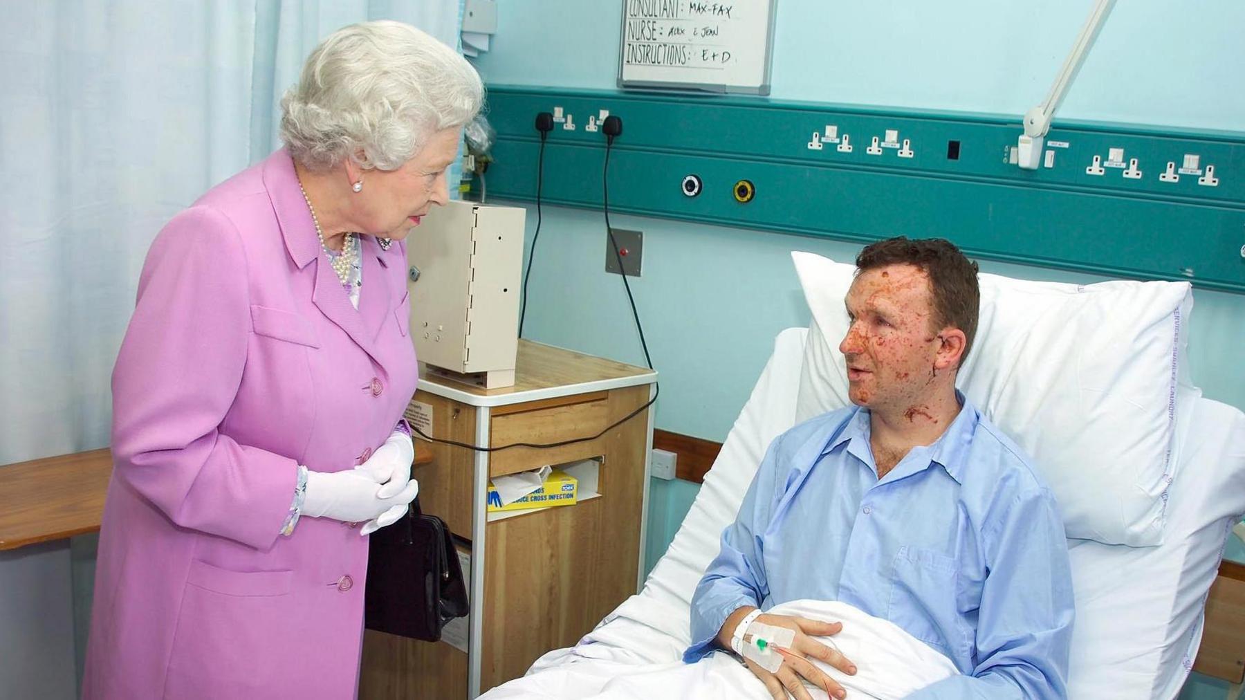 Mr Lait in a hospital bed as he speaks with Queen Elizabeth II. He is propped up on white pillows and is wearing blue pyjamas. His face is scarred. The Queen is wearing a pink dress and, as she stands by the bed, she is leaning forward slightly as she speaks to Mr Lait.