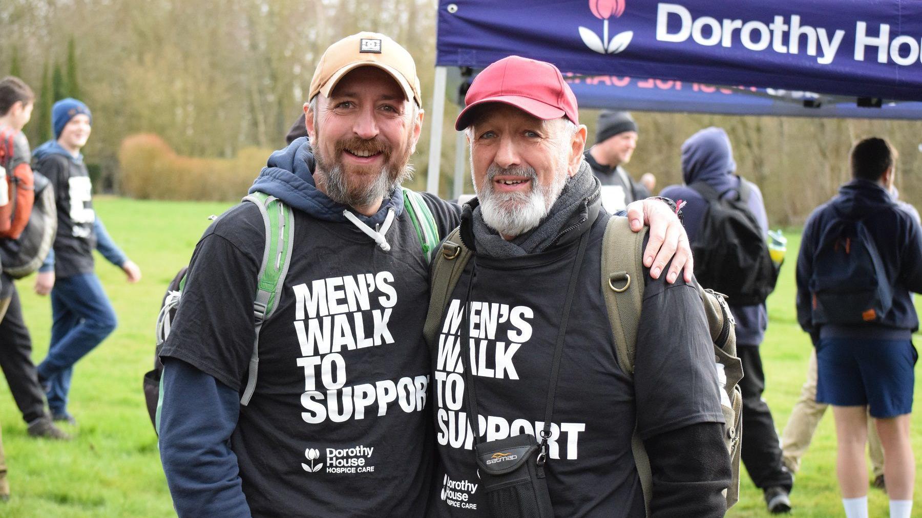 Two men stand together on a field. They both are wearing caps, backpacks and black T-shirts which say 'Men's walk to support' over long-sleeved jumpers. They are standing in front of people who are gathered by a purple gazebo which says 'Dorothy House'.