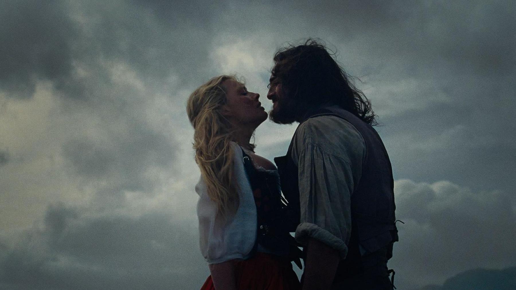 Film still with Margot Robbie and Jacob Elordi outside under a glowering, cloud-filled sky. The light is low as if a storm is imminent, and the wind is blowing both of their hair as they stand facing each other.