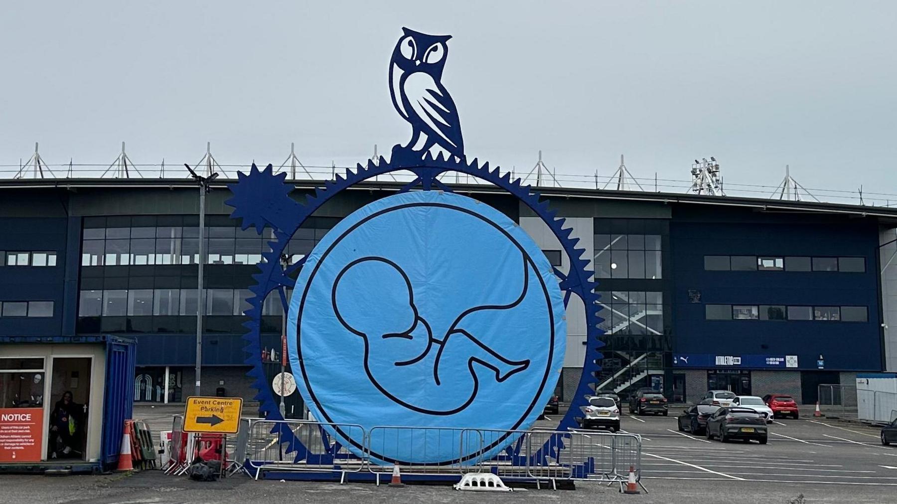 Line drawing of embryonic baby in a blue circle, which is draped inside a sculpture of a cog with the owl mascot of Oldham Athletic above. The work appears outside the car park in front of the stadium.