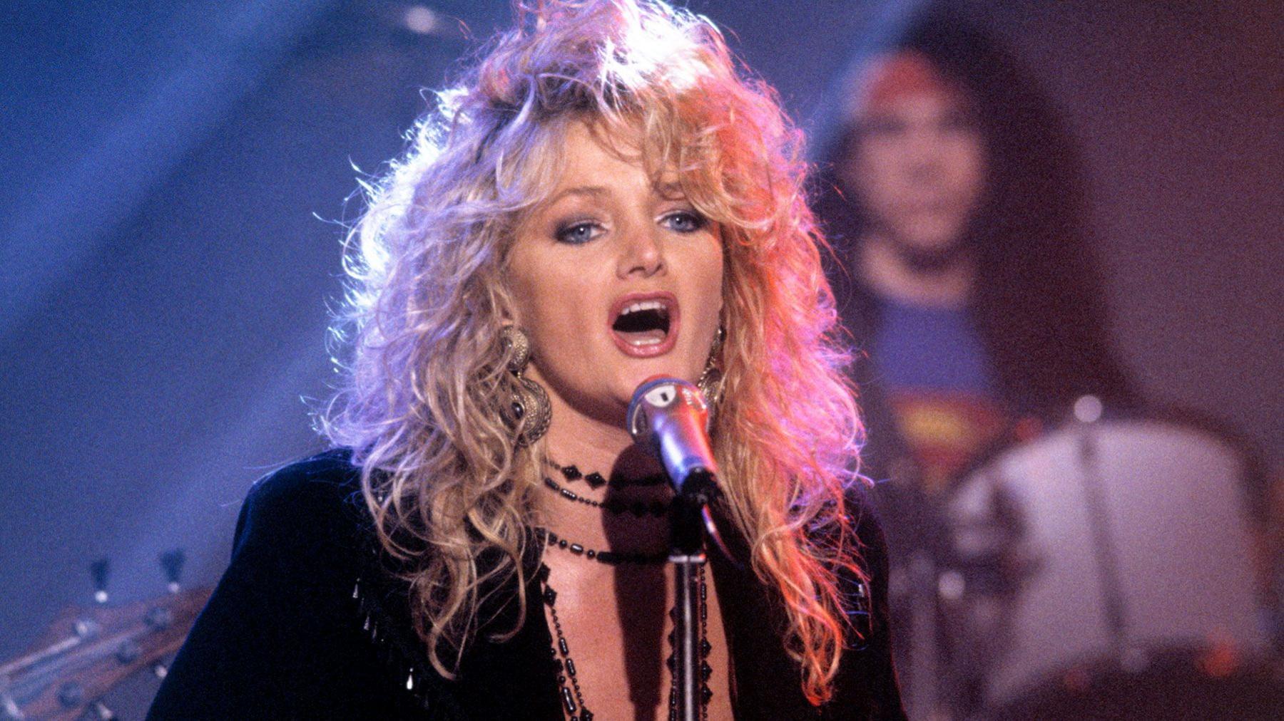 Bonnie Tyler singing into a microphone. She has big blonde hair and a black dress on. Behind is a drummer - he is slightly blurred, as Bonnie is the focus of the image.