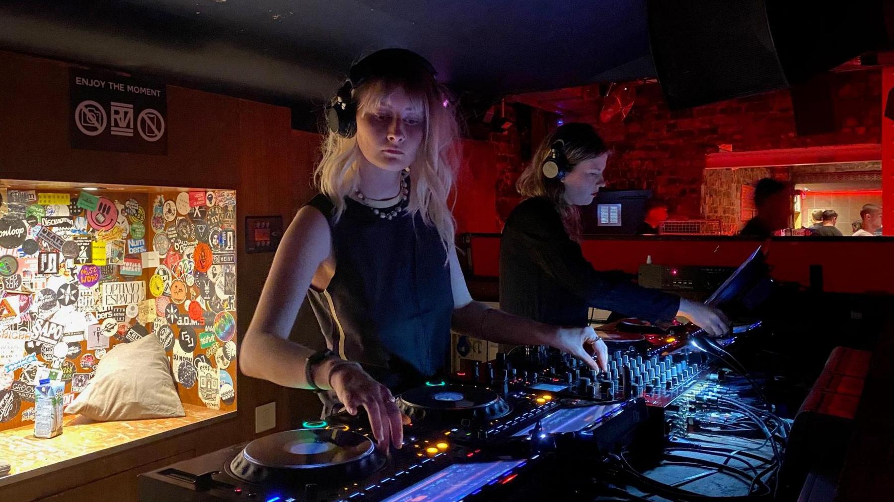 Two women DJ on a set of decks in a dark club.