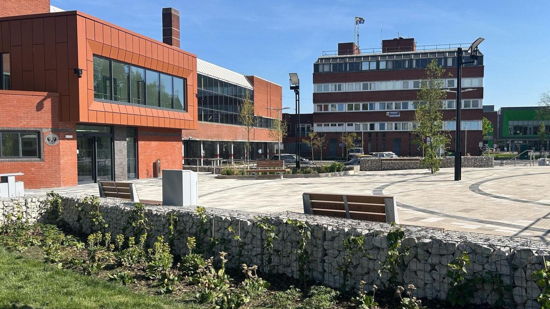 A paved open space, with trees and lighting columns as well as a wall which has planting in front of it and benches to one side. The space is bordered by tall buildings. There are further benches and planting located around the paved area.